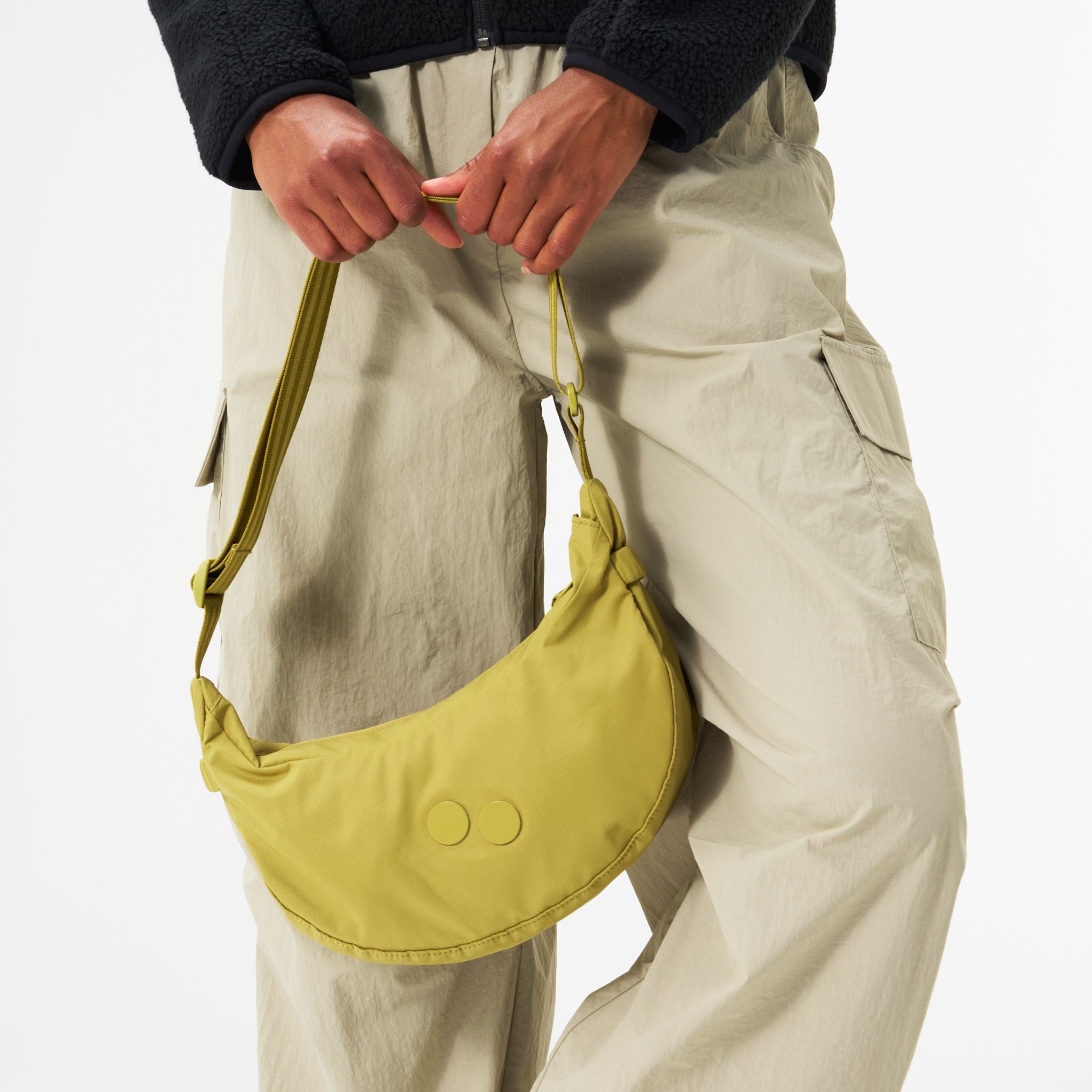 Person hält eine gelbe Umhängetasche, trägt beige Cargohosen und eine schwarze Jacke.