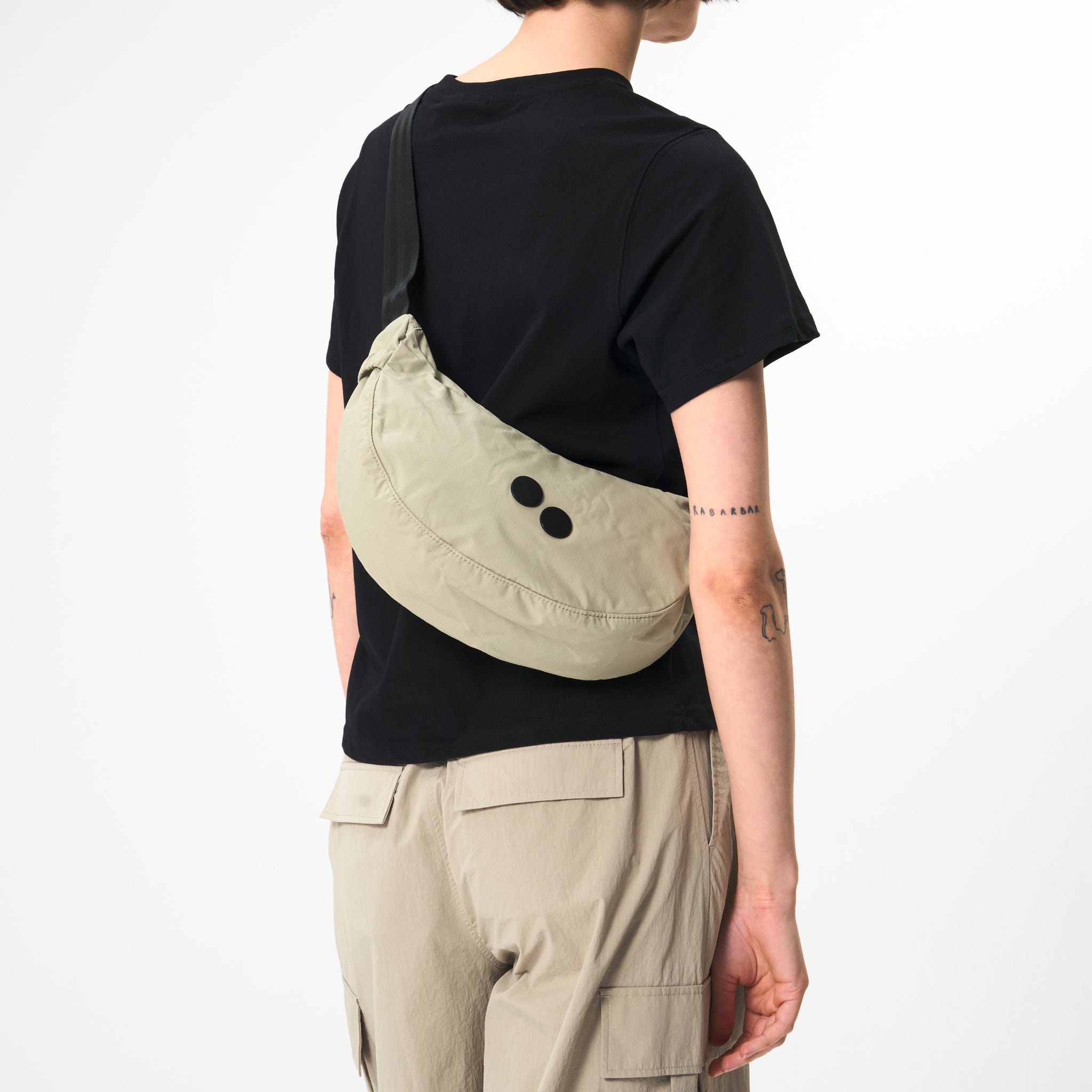 Person in schwarzem T-Shirt trägt eine beige Umhängetasche und beige Hose mit Taschen, mit Tattoos auf dem Arm.