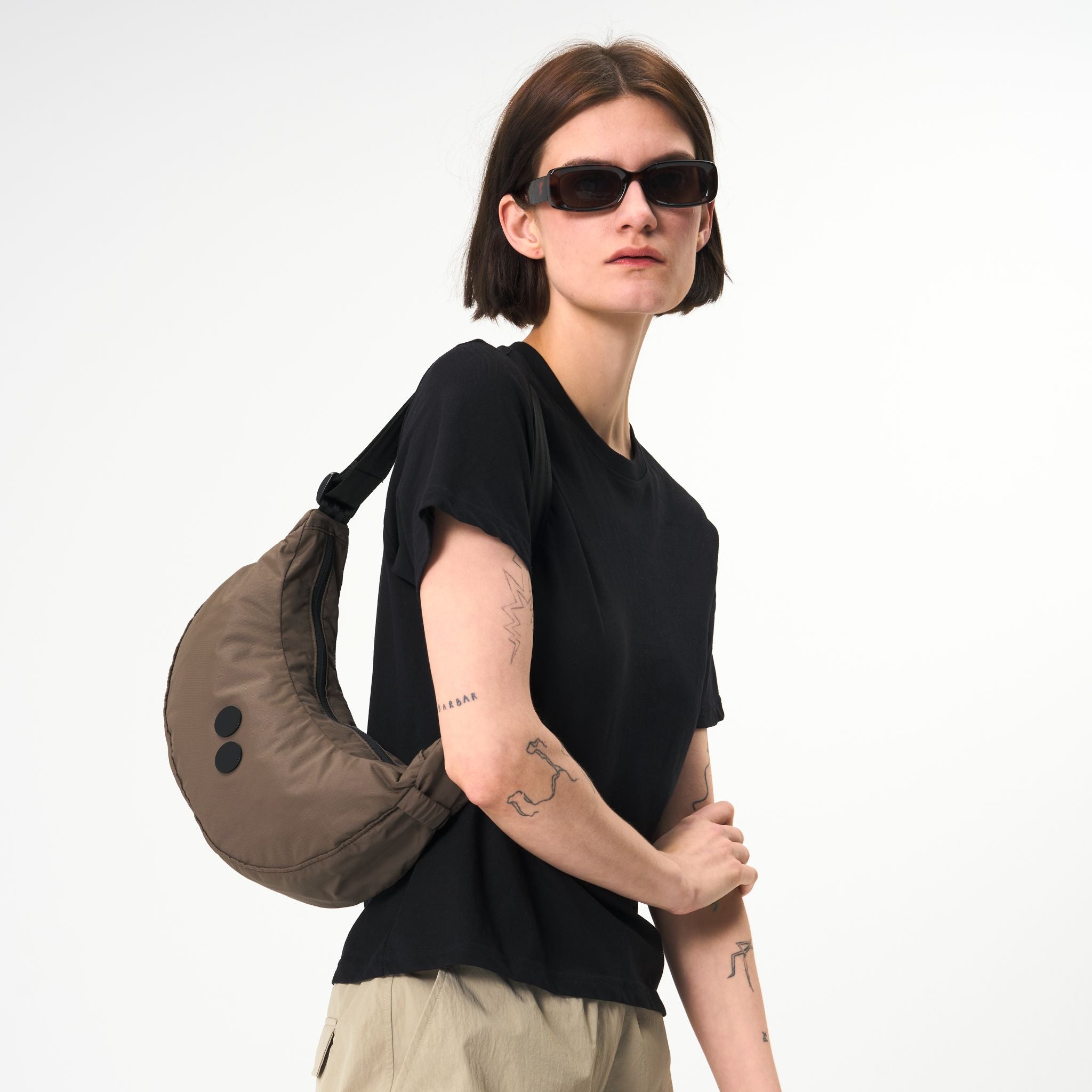 Person mit Sonnenbrille, schwarzem T-Shirt und beiger Umhängetasche vor weißem Hintergrund.