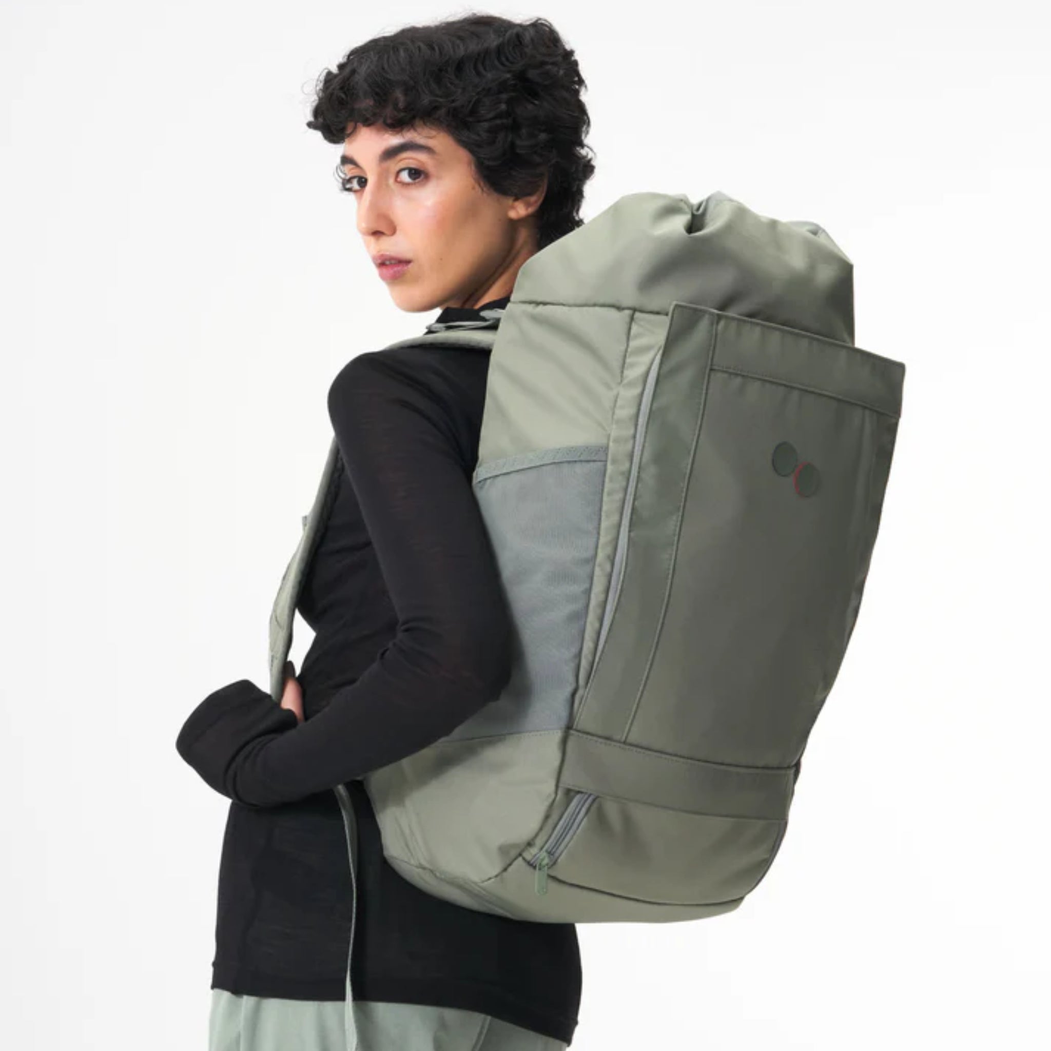 Eine Person mit kurzem, lockigem, dunklem Haar trägt einen großen, hellgrünen Rucksack und ein schwarzes, langärmeliges Oberteil. Sie steht seitlich und blickt über die Schulter vor einem einfarbig weißen Hintergrund zurück.