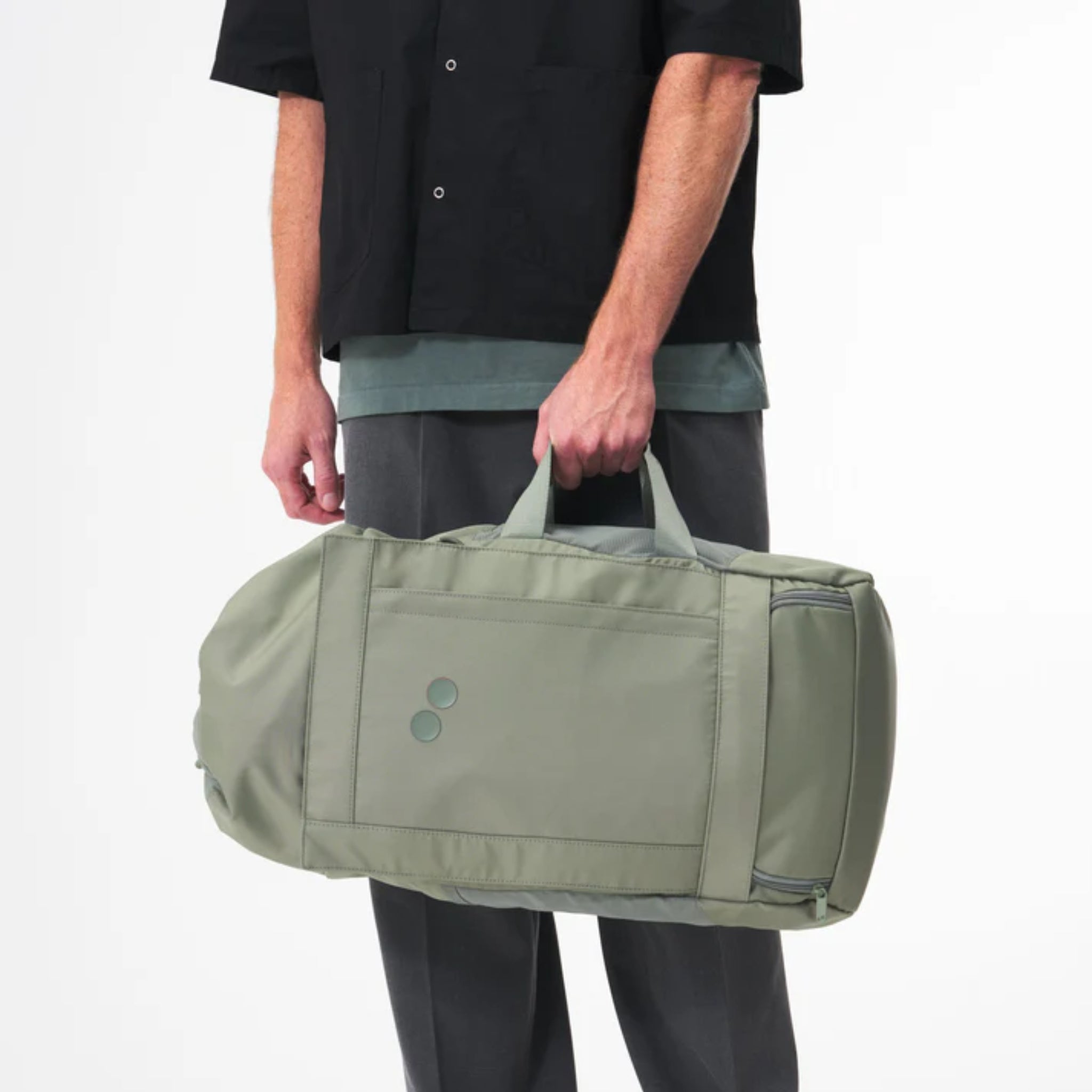 Eine Person, die ein schwarzes kurzärmeliges Hemd und eine graue Hose trägt, hält einen hellgrünen Seesack an den Griffen vor einem schlichten weißen Hintergrund.