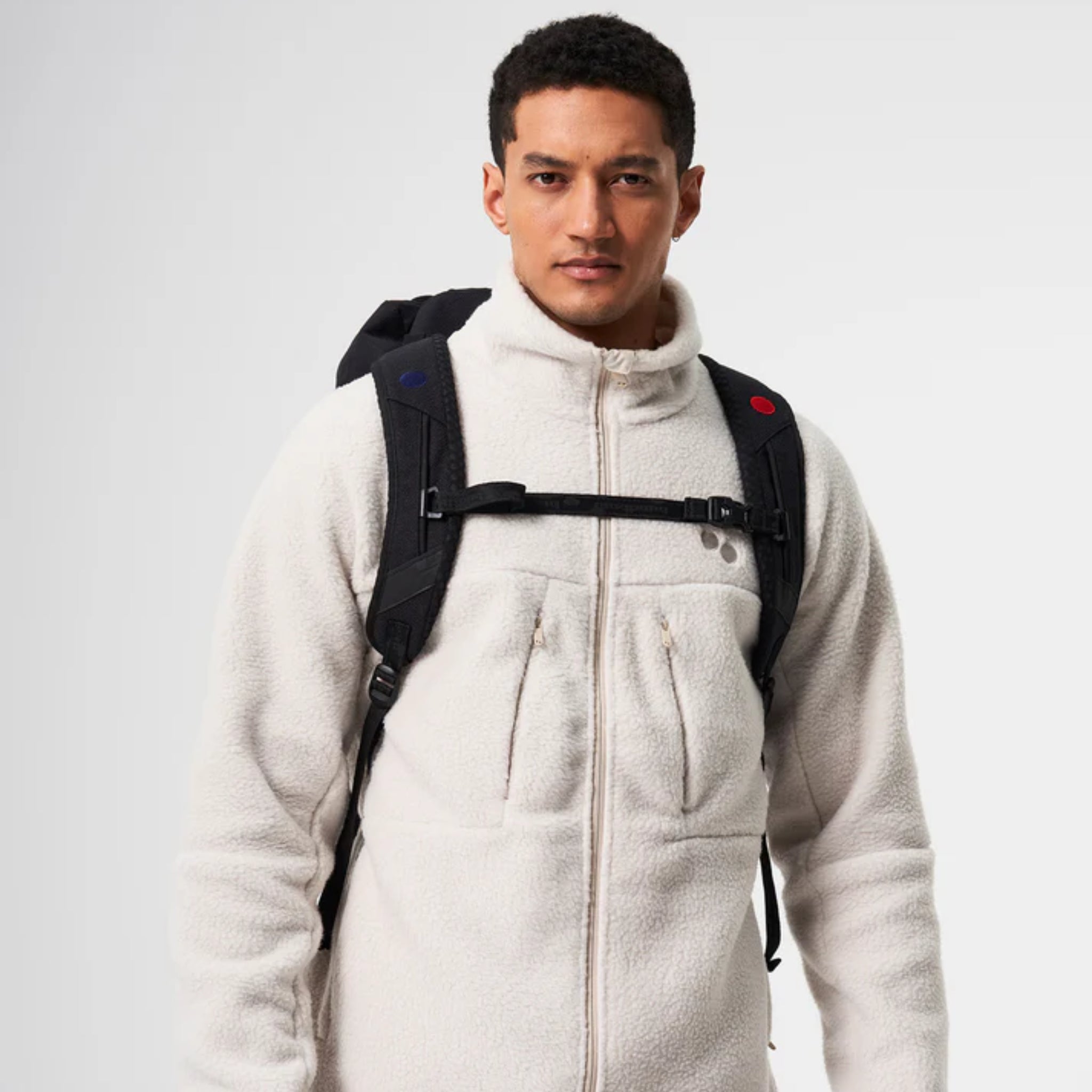 Ein Mann, der eine weiße Fleecejacke und einen schwarzen Rucksack trägt, steht mit dem Gesicht nach vorne vor einem schlichten hellgrauen Hintergrund.