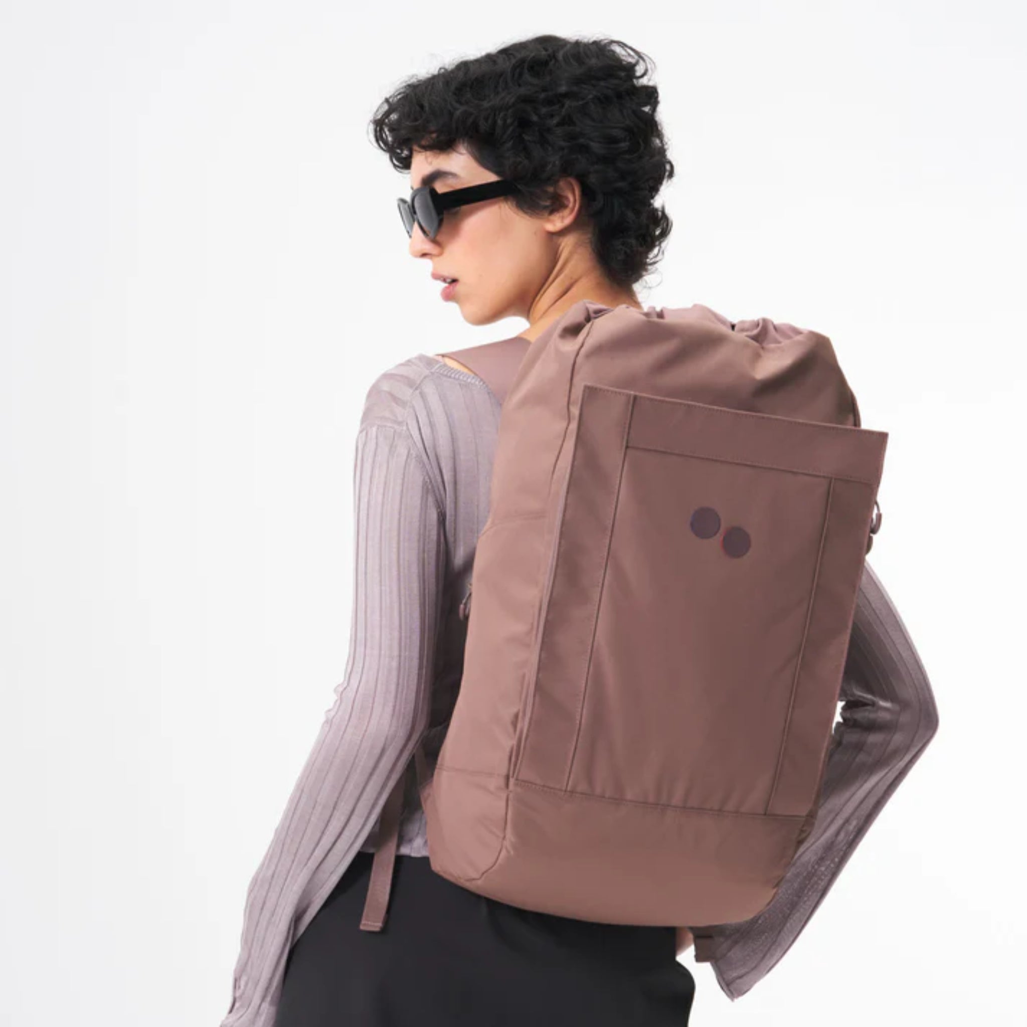 Eine Person mit kurzen lockigen Haaren, schwarzer Sonnenbrille und einem hellen, langärmeligen Oberteil steht seitlich und modelliert einen großen lilafarbenen Rucksack mit zwei kleinen dunklen Kreisen auf der Gesäßtasche vor einem schlichten weißen Hintergrund.
