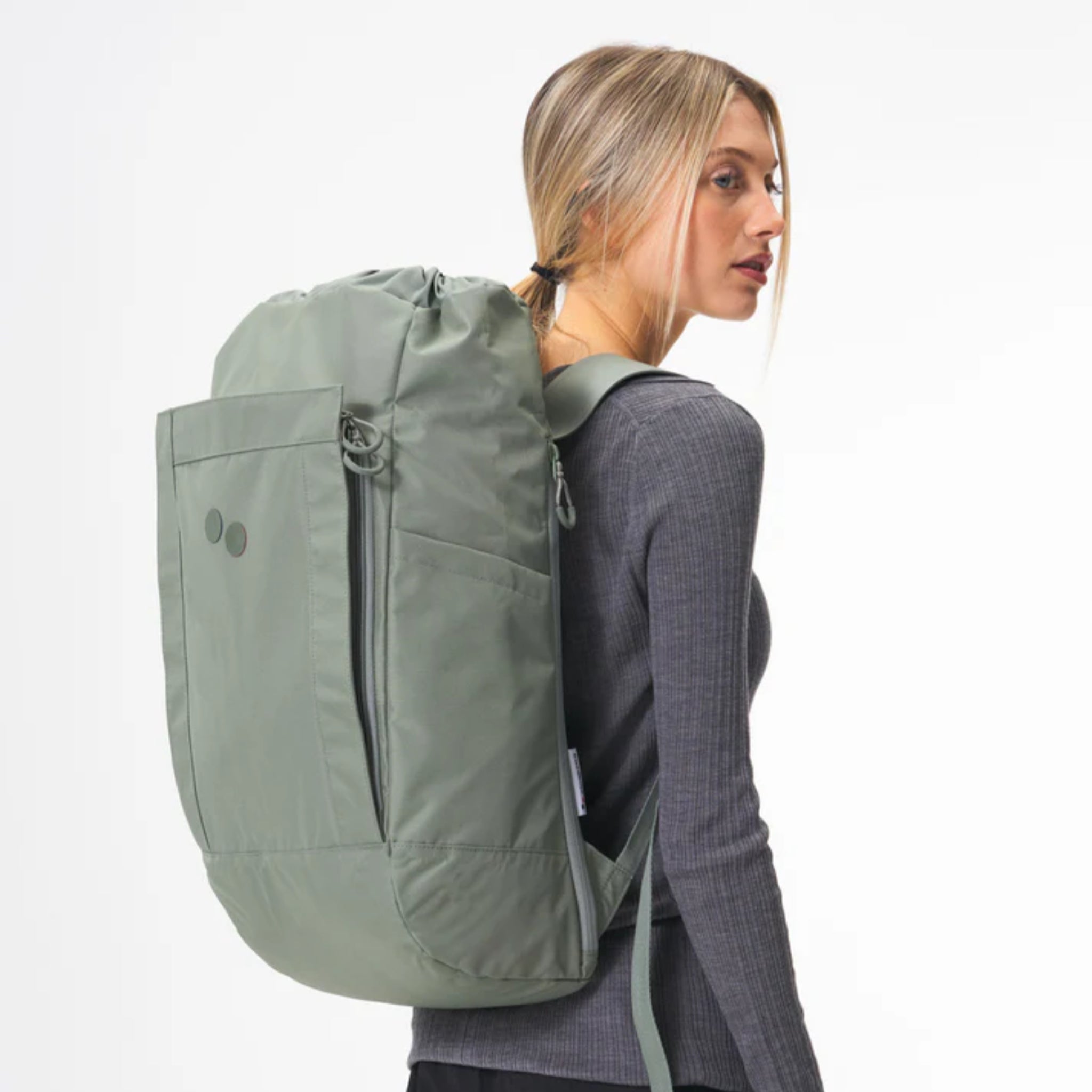 Eine Frau mit blondem Haar, die ein langärmeliges graues geripptes Hemd trägt, trägt einen großen hellgrünen Rucksack über der Schulter und blickt nach rechts vor einem schlichten, hellen Hintergrund.