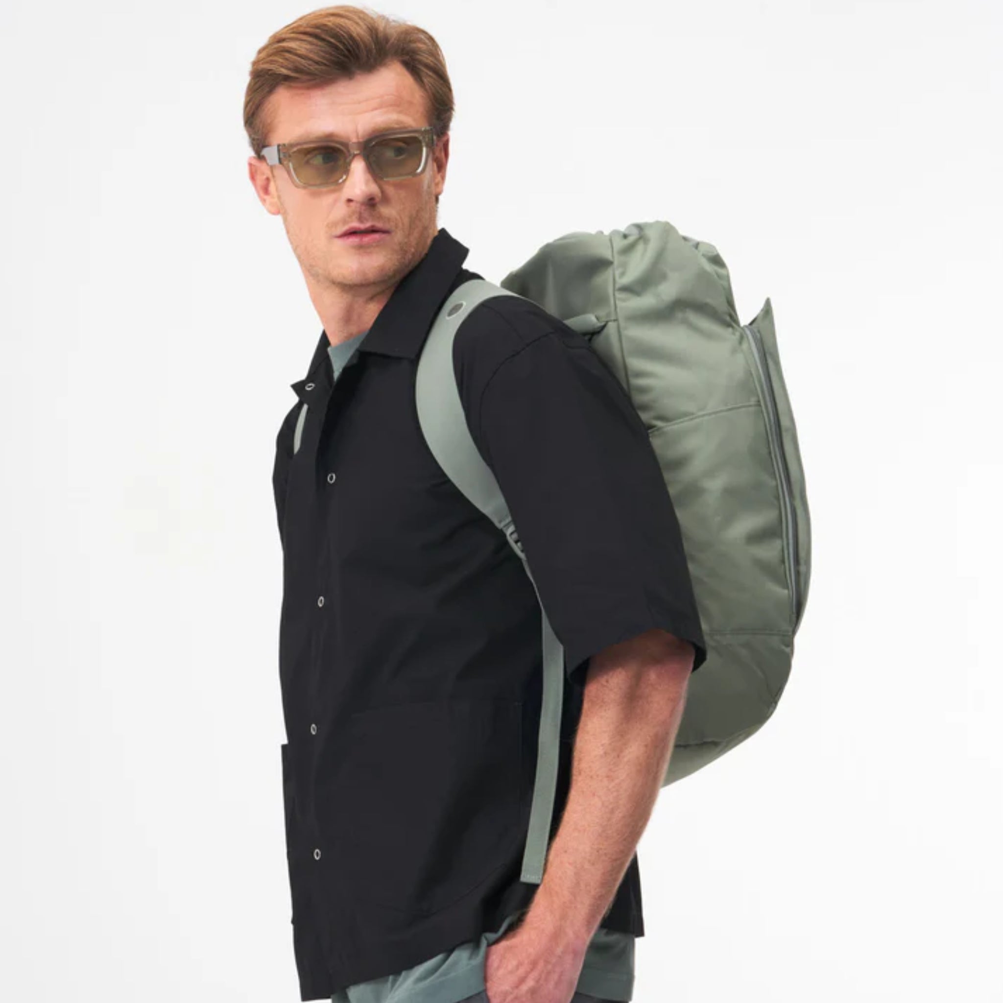 Ein Mann mit hellbraunem Haar, der eine Sonnenbrille, ein schwarzes, kurzärmeliges Button-up-Hemd und einen grünen Rucksack über einer Schulter trägt, blickt vor einem schlichten weißen Hintergrund zur Seite.