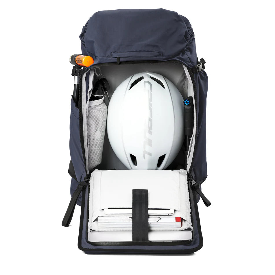 Ein dunkelblauer Rucksack wird vollständig geöffnet und gibt den Blick auf ein ordentlich gepacktes Inneres frei. Zu sehen sind ein weißer Helm, ein zusammengerolltes Handtuch, eine Taschenlampe, eine Wasserflasche und geordnete weiße Ordner oder Aktenordner.