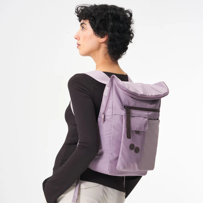 Eine Person mit kurzen, lockigen dunklen Haaren, die ein langärmeliges schwarzes Oberteil und eine helle Hose trägt, steht seitlich und trägt einen hellvioletten Rucksack mit Vordertaschen vor einem schlichten weißen Hintergrund.