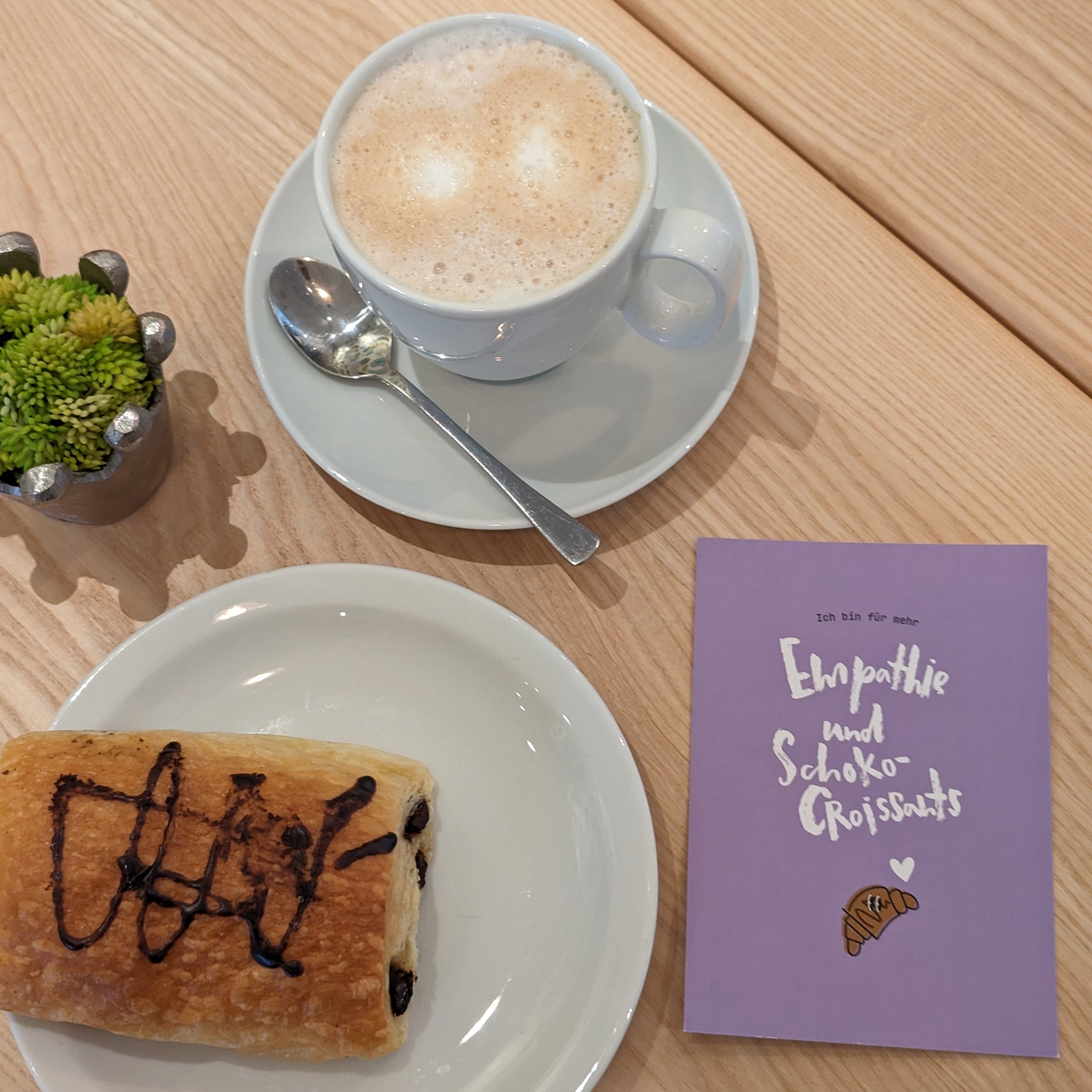 Tasse Cappuccino, Schokocroissant auf einem Teller und eine lilafarbene Karte mit der Aufschrift "Empathie und Schoko-Croissants".