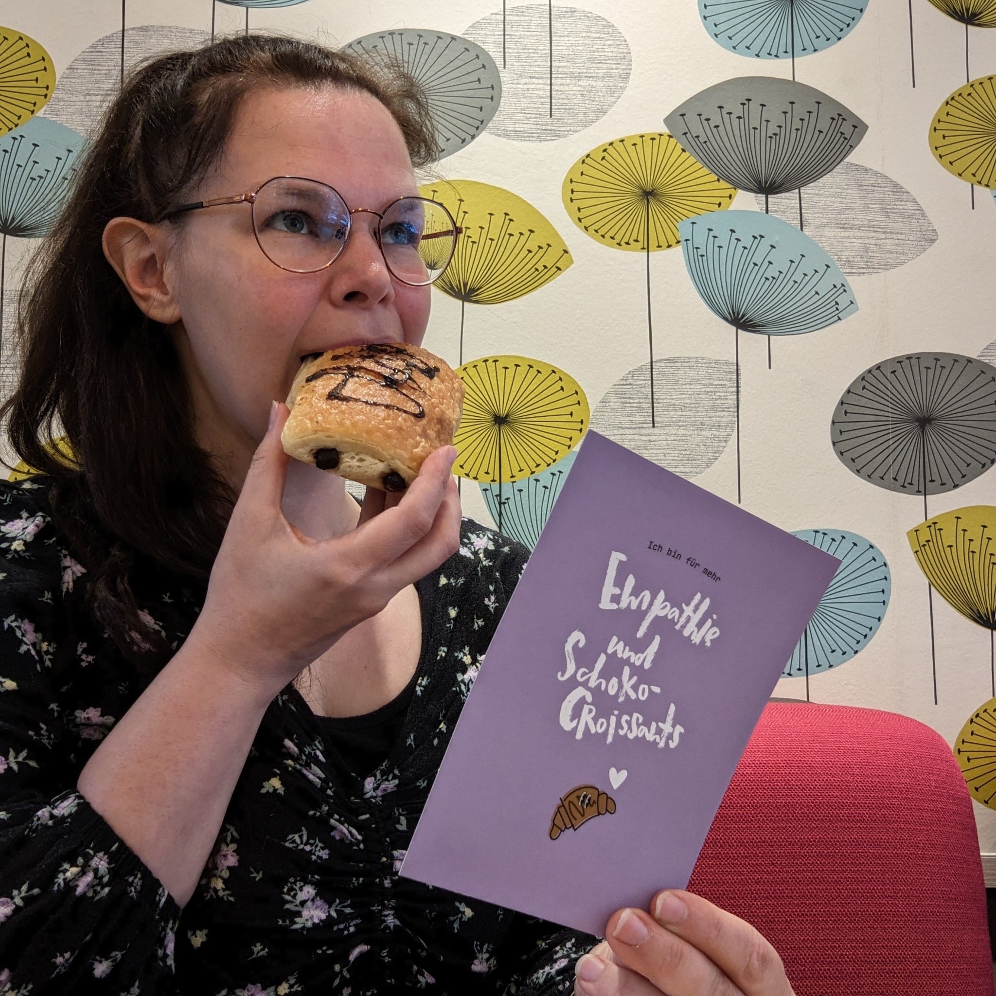 Frau isst ein Schokocroissant und hält eine Karte mit der Aufschrift "Empathie und Schoko-Croissants" in einem bunten Raum.