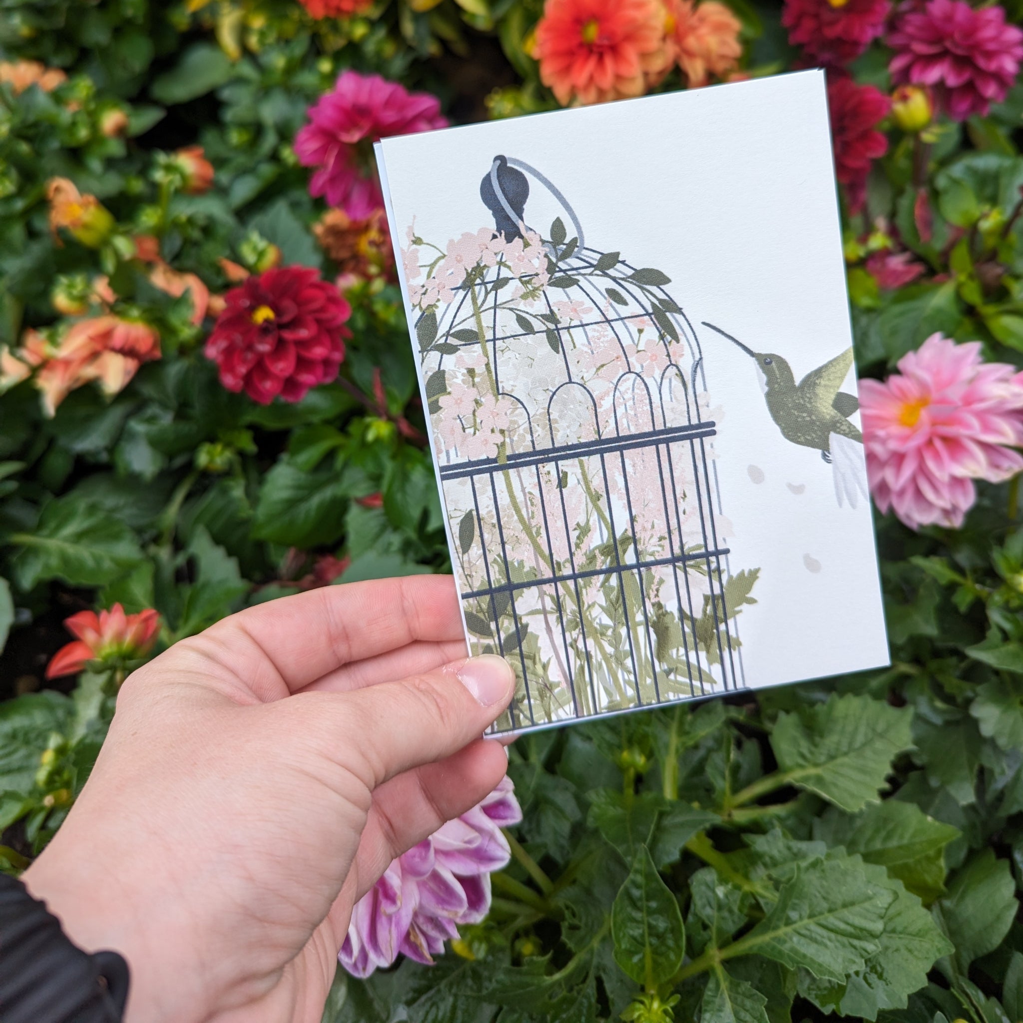 Eine Hand hält eine Karte mit einem Vogelkäfig und einem Kolibri, umgeben von bunten Blumen.