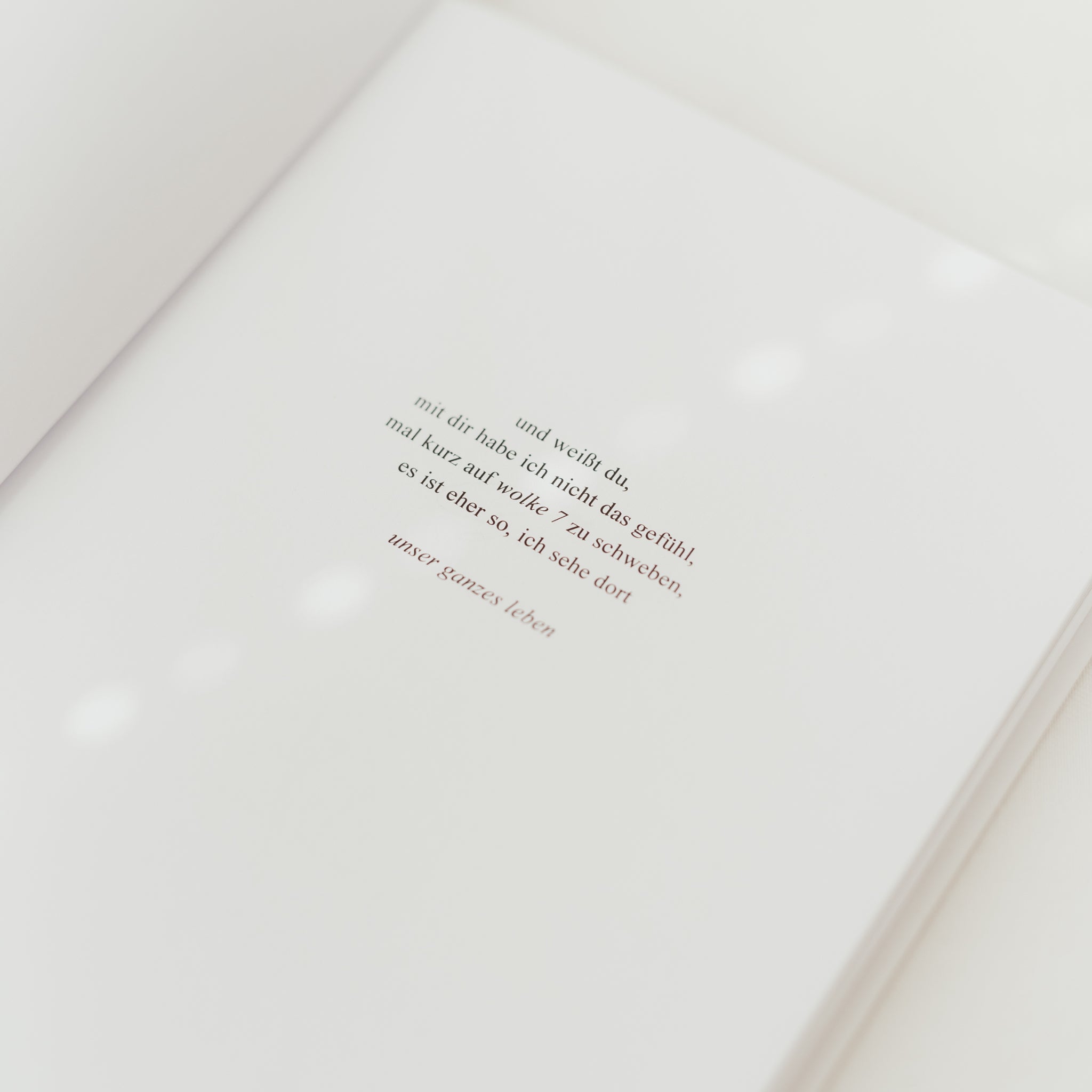 Auf der aufgeklappten Seite eines Buches steht ein poetischer Text in schwarzer und roter Schrift auf weißem Hintergrund.