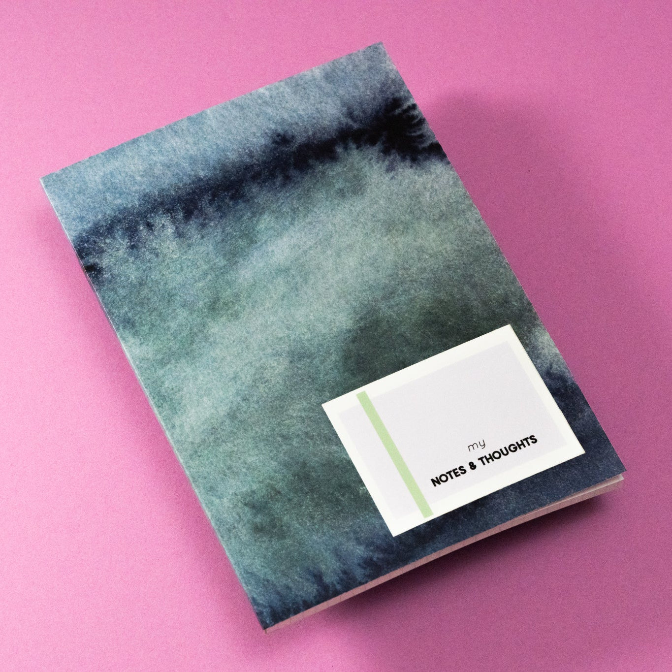 Ein Notizbuch mit blauem und grünem Einband im Aquarellstil liegt auf einem rosa Hintergrund. Obenauf liegt eine kleine weiße Karte, auf der der Text my NOTES & THOUGHTS aufgedruckt ist.