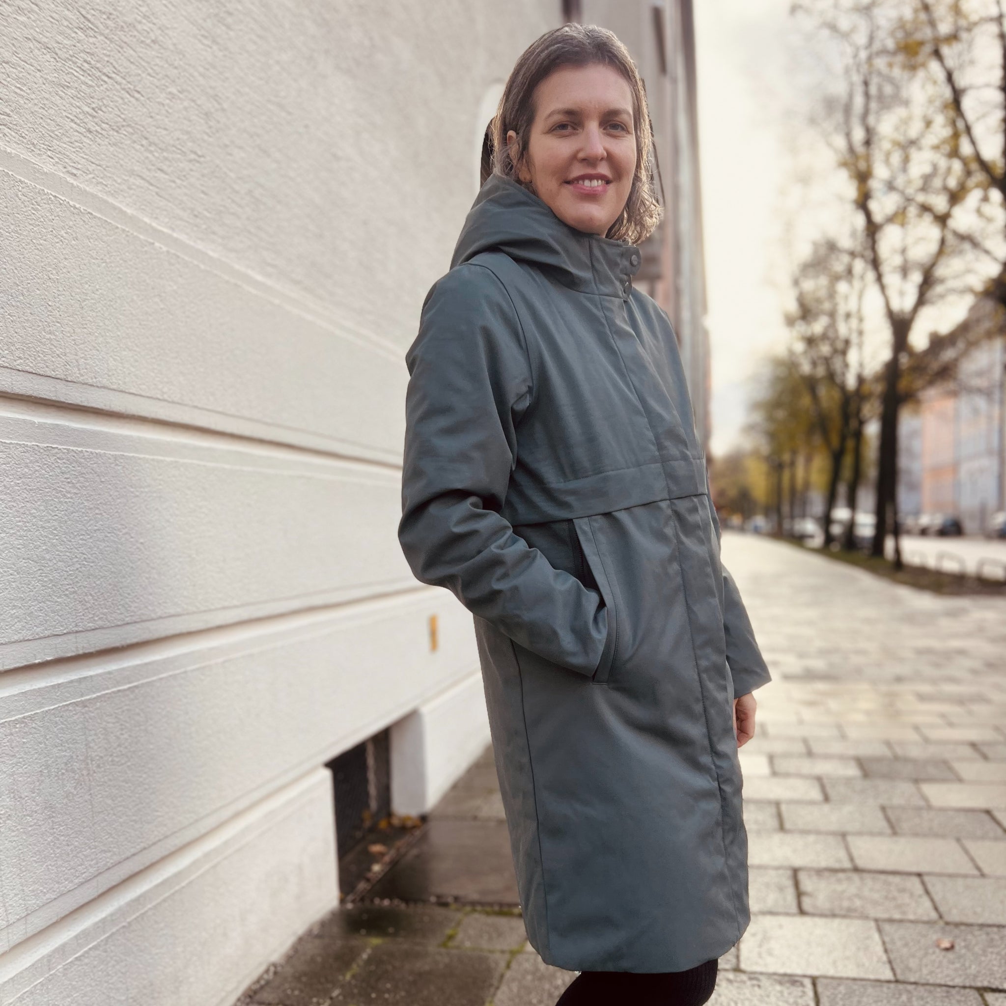 Eine Person in einem grauen Mantel steht auf einem Bürgersteig neben einer weißen Wand an einem bewölkten Tag.