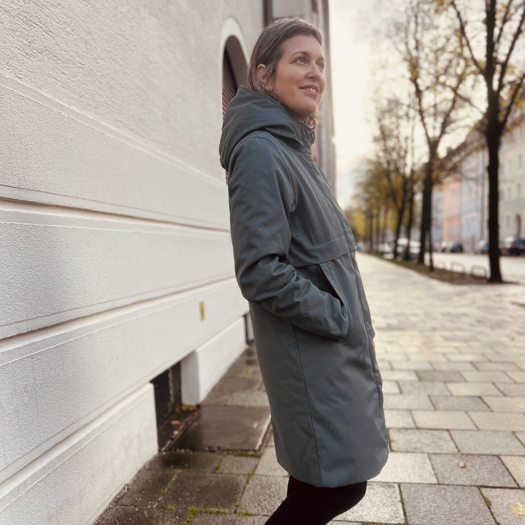 Eine Frau steht in einem grauen Mantel auf einem Bürgersteig vor einer weißen Wand, umgeben von Bäumen.
