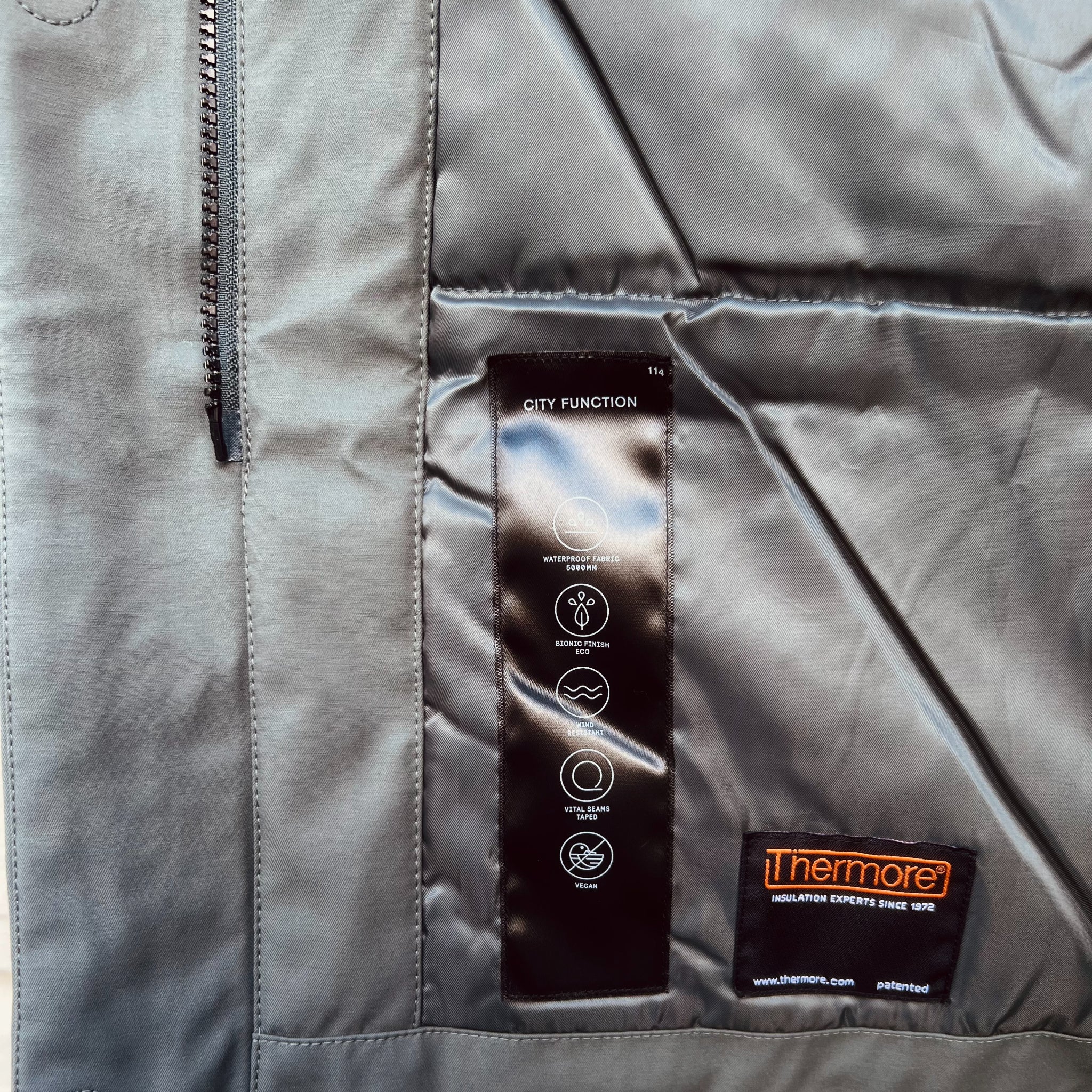 Graue Jacke mit schwarzem Etikett, das Funktionen wie wasserfest, vegan und isolierend zeigt.