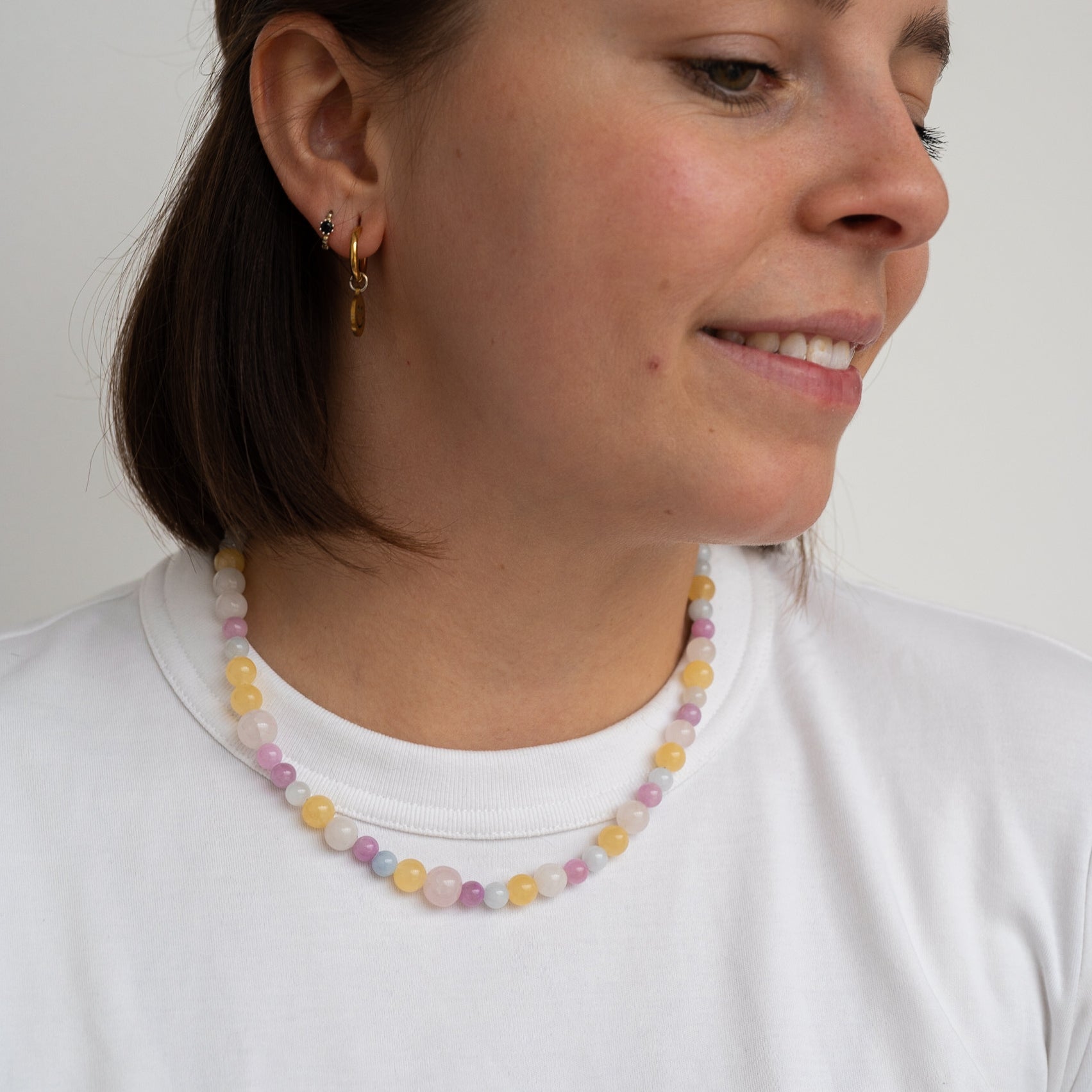 Eine Frau, die ein weißes T-Shirt und eine Perlenkette mit pastellfarbenen Perlen sowie kleine goldene Ohrringe trägt, lächelt und schaut leicht zur Seite.