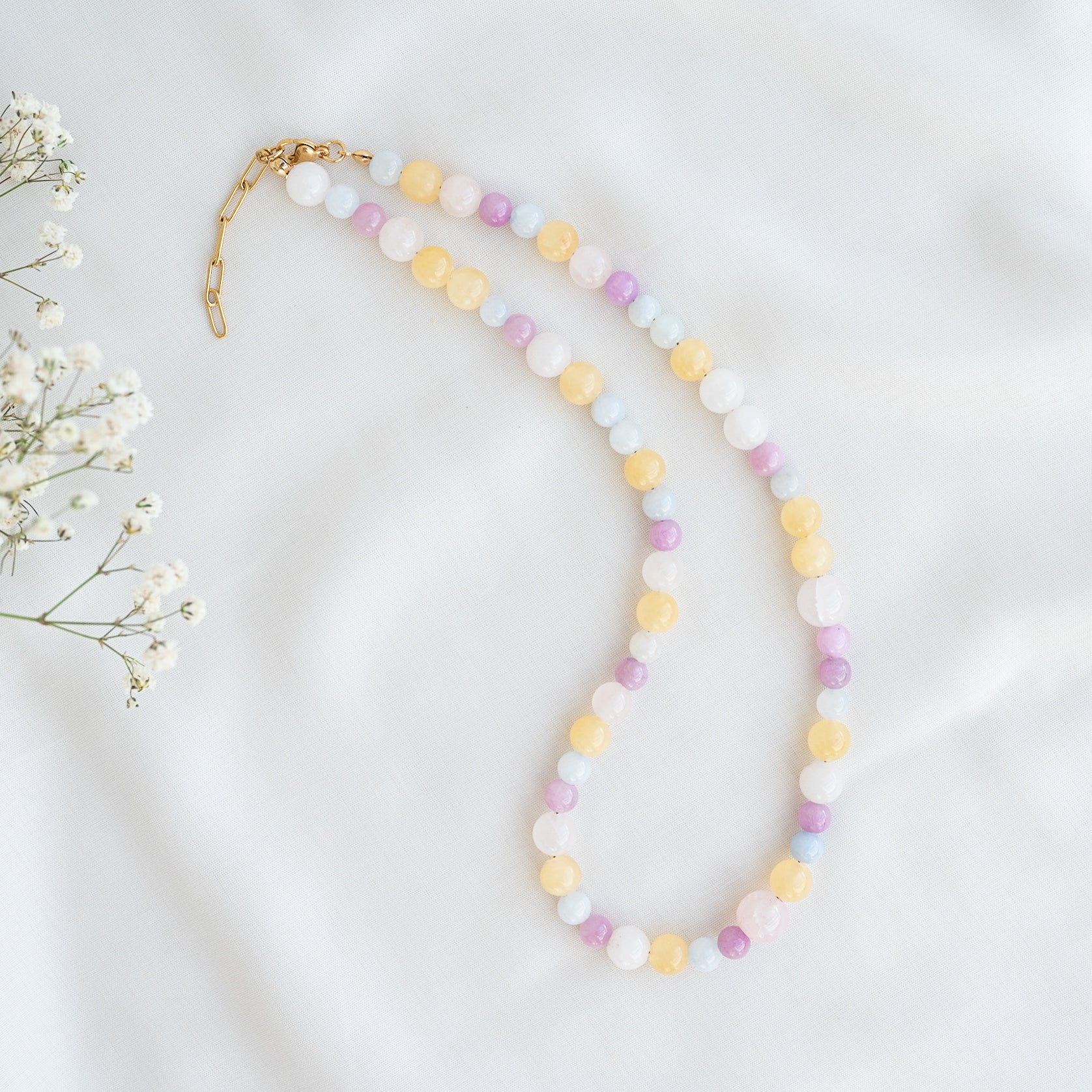 Eine Perlenkette mit pastellgelben, lila, blauen und weißen runden Perlen ist auf einem weißen Stoff ausgelegt. Ein goldener Kettenverschluss ist angebracht. Auf der linken Seite sind Baby's Breath-Blüten zu sehen.
