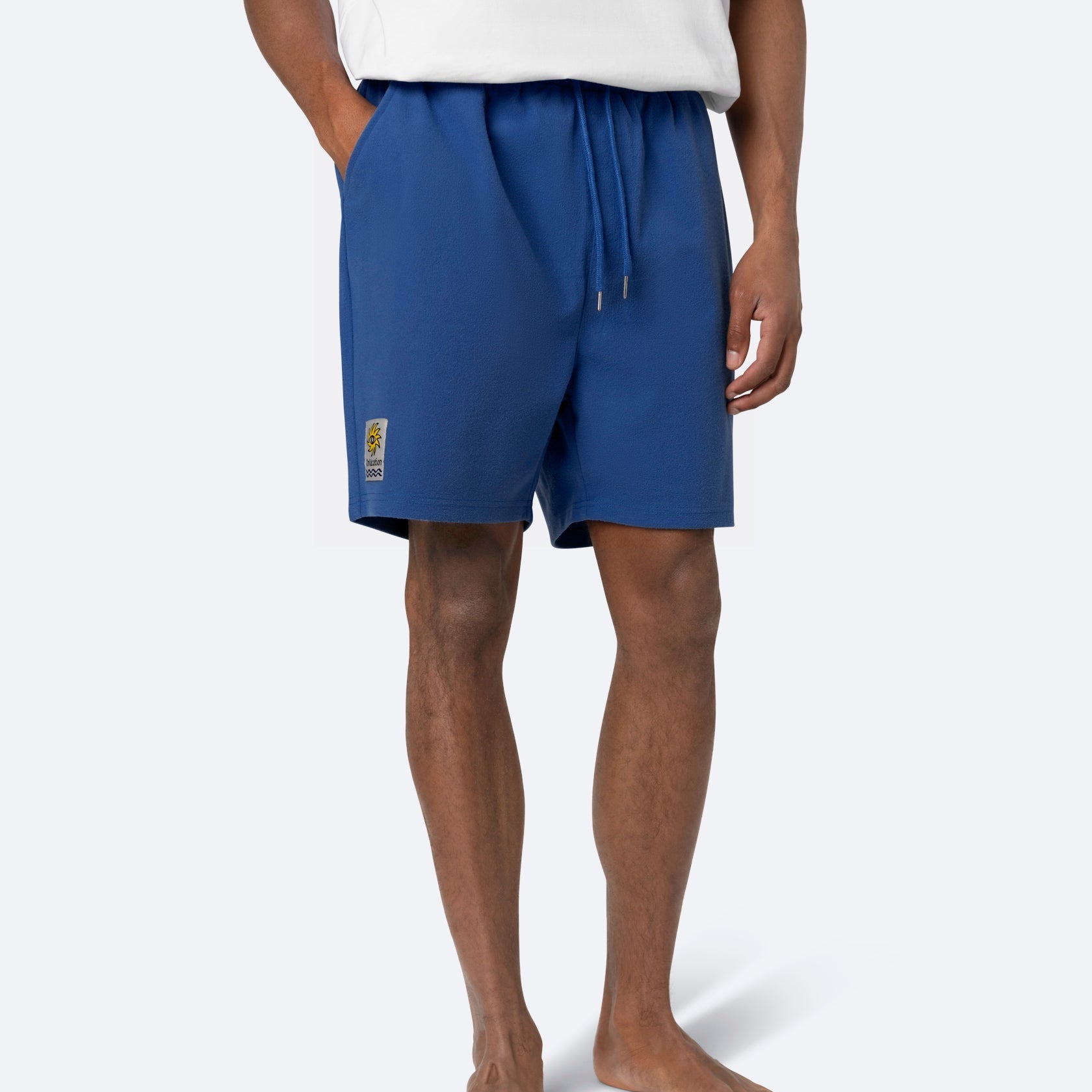 Eine Person trägt ein weißes Hemd und blaue Shorts mit Kordelzug, die Hände in den Taschen. Die Shorts haben einen kleinen gelben Aufnäher am linken Bein. Der Hintergrund ist einfarbig und hell gefärbt.