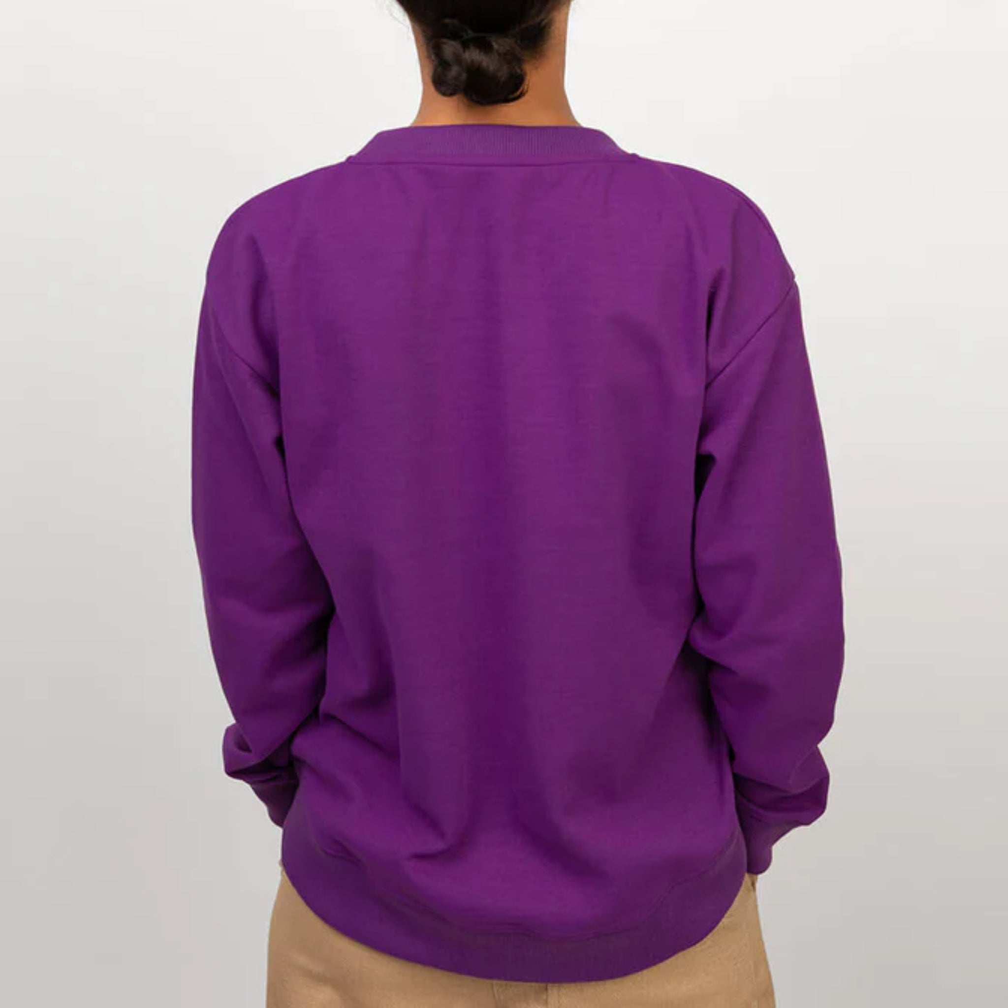 Eine Person steht mit dem Gesicht zur Kamera, trägt ein hellviolettes langärmeliges Sweatshirt und eine hellbraune Hose vor einem schlichten weißen Hintergrund.