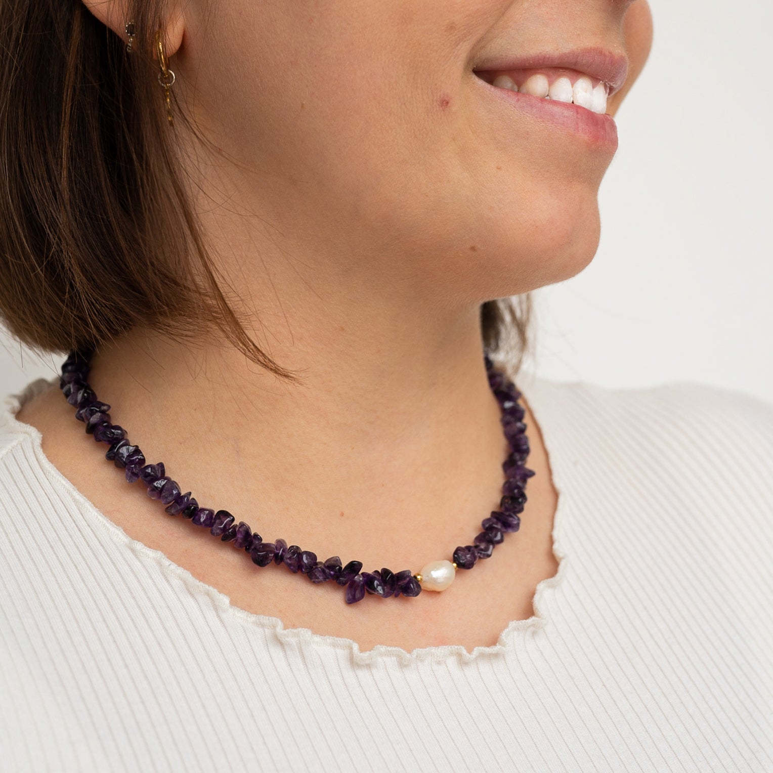 Eine Frau, die eine violette Edelsteinkette mit einer einzelnen weißen Perle trägt, lächelt und ist mit einem weißen gerippten Oberteil mit gerafftem Ausschnitt bekleidet.
