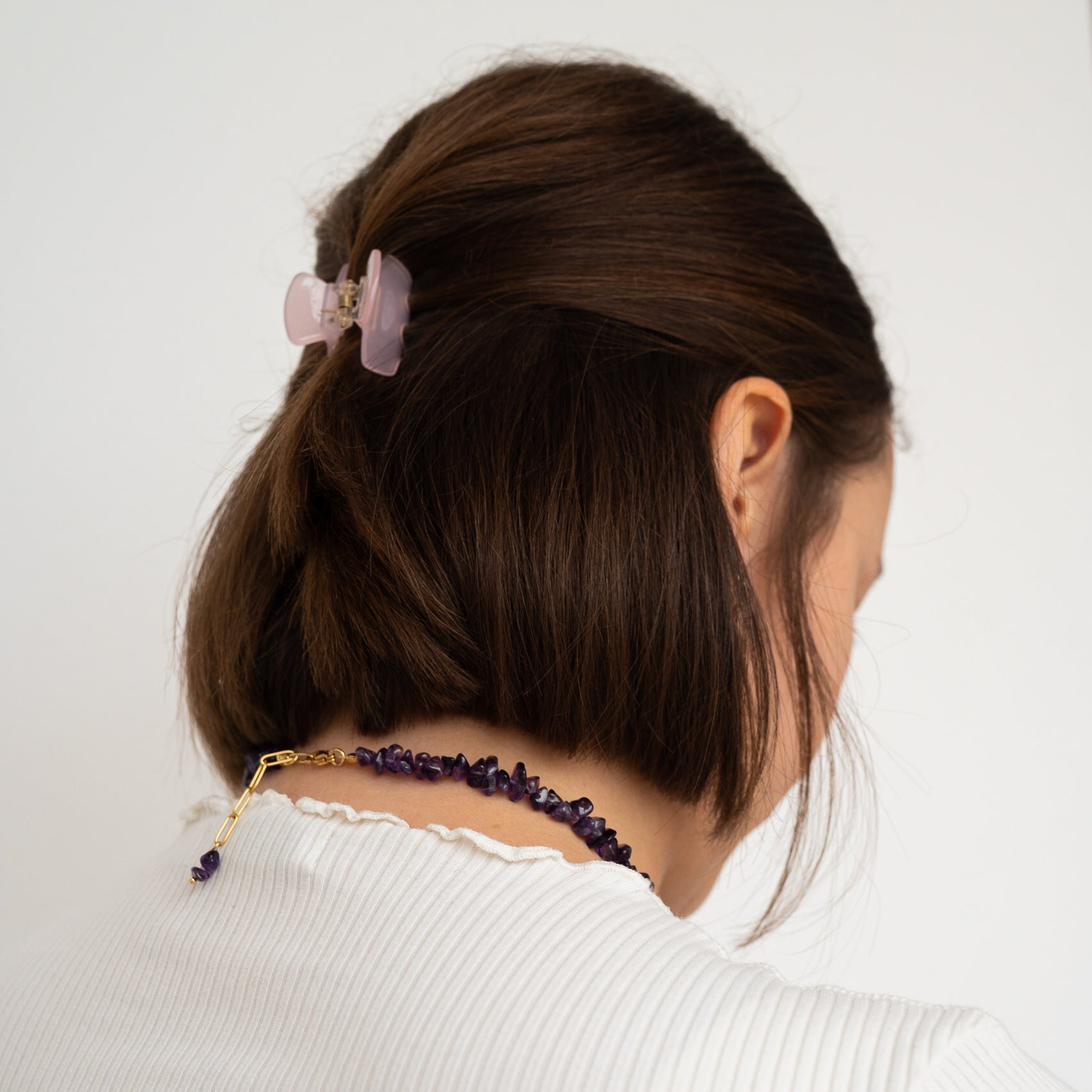 Eine Frau mit braunem Haar, das mit einer hellrosa Haarspange hochgesteckt ist, wird von hinten gezeigt. Sie trägt ein weißes Oberteil mit gerüschtem Ausschnitt und eine Halskette aus lila Steinen.