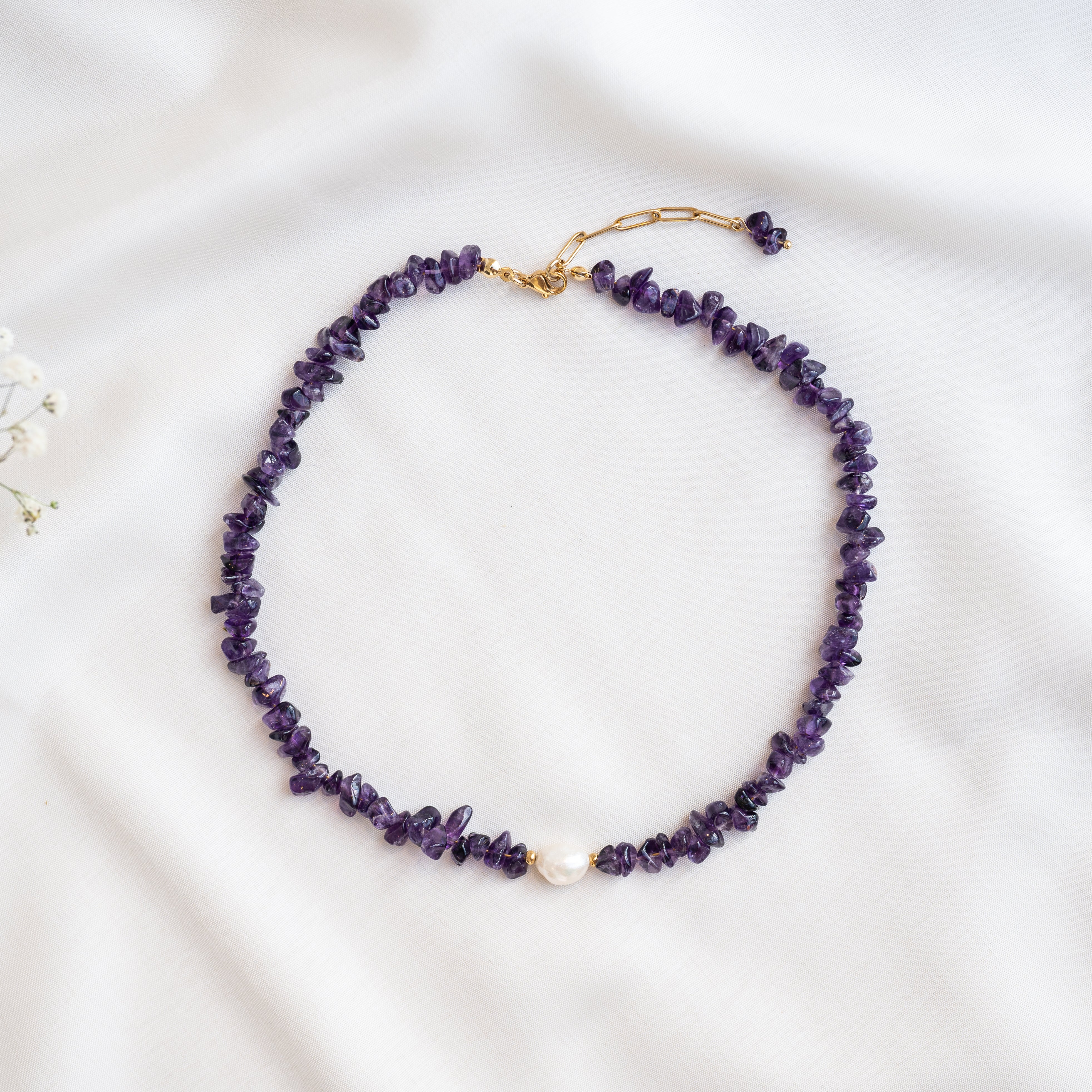 Eine Halskette aus unregelmäßigen violetten Edelsteinperlen mit einer einzelnen weißen Perle in der Mitte, die auf einem weißen Stoffhintergrund angebracht ist. Die Halskette hat einen goldenen Verschluss und eine verstellbare Kette.