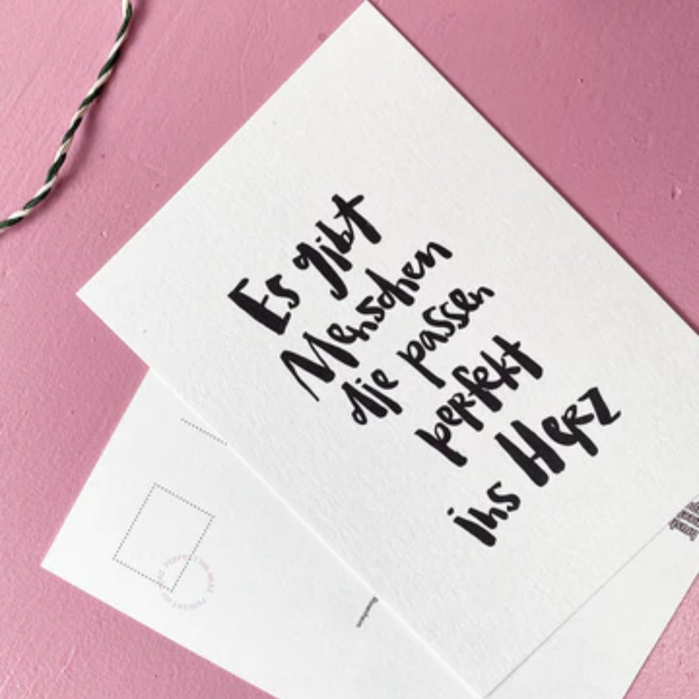 Auf einer weißen Postkarte mit deutscher Handschrift steht "Es gibt Menschen die perfekt ins Herz passen" auf einer rosafarbenen Fläche, daneben eine weitere einfarbige Postkarte und eine schwarz-weiße Schnur.