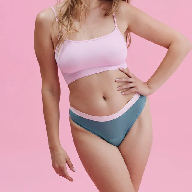 Eine Frau posiert selbstbewusst vor einem rosafarbenen Hintergrund. Sie trägt ein hellrosa Crop-Top und hochtaillierte, blaugrüne Unterwäsche mit einem rosafarbenen Bund. Sie hat langes, gewelltes Haar und buntes Augen-Make-up.