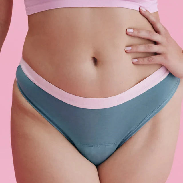 Eine Person, die blaue Unterwäsche mit einem hellrosa Bund und ein dazu passendes rosafarbenes Crop-Top trägt, steht vor einem rosafarbenen Hintergrund und stützt eine Hand auf ihre Hüfte.