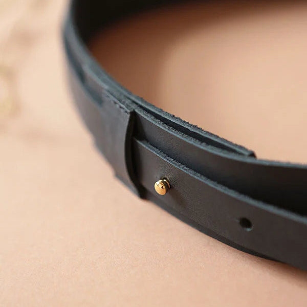 Nahaufnahme eines schwarzen Ledergürtels mit einem kleinen goldenen Nietenbesatz auf einer hellbeigen Oberfläche. Der Gürtel ist teilweise geschlossen, und die Textur des Leders ist sichtbar.