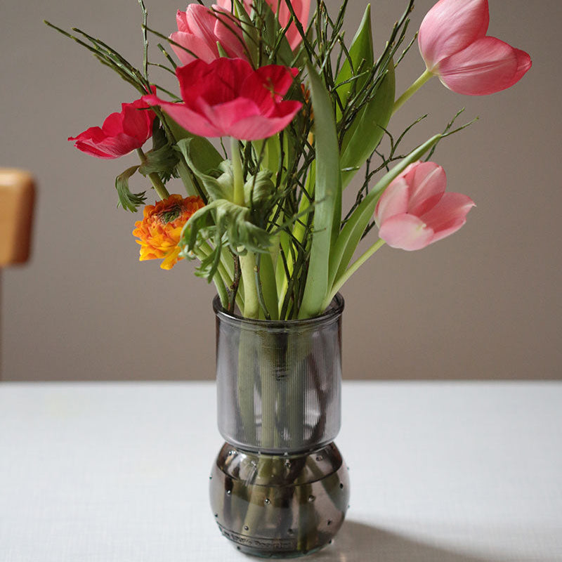 Ein Strauß roter, rosafarbener und orangefarbener Blumen mit grünen Stielen, arrangiert in einer modernen, zweiteiligen Vase aus dunklem Glas auf einem hellen Tisch.