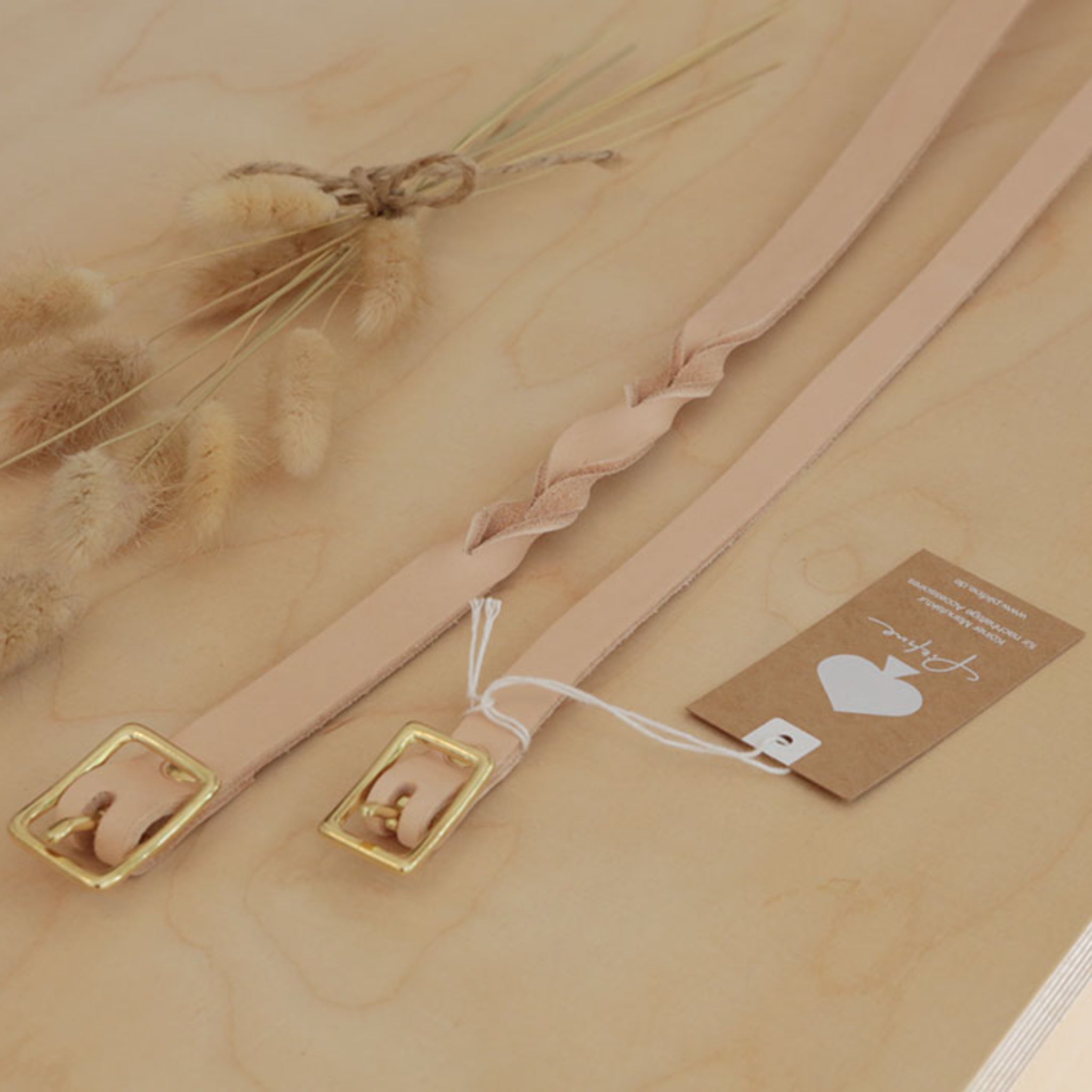 Zwei beigefarbene Lederriemen mit goldenen Schnallen liegen auf einem Tisch neben einem Strauß trockener Gräser.