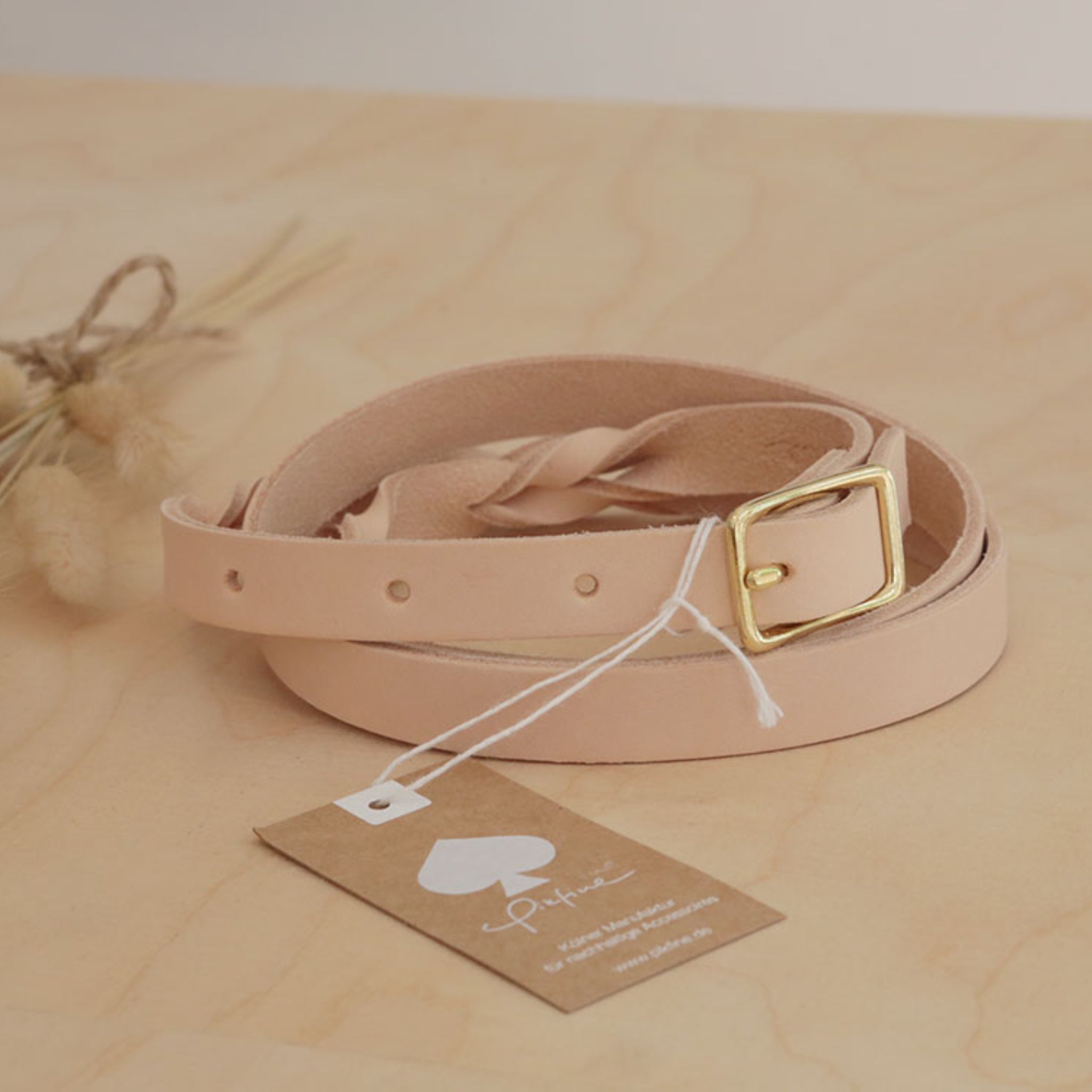 Hellbrauner Ledergürtel mit goldener Schnalle liegt auf einem Holztisch. Daneben ein Etikett und Deko Gräser.