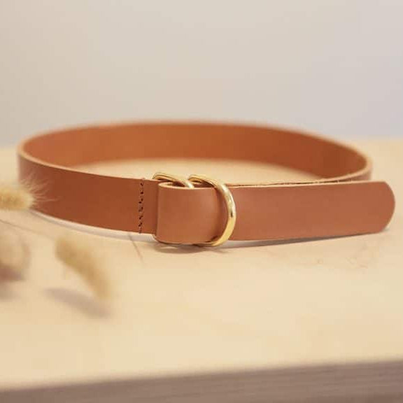 Ein hellbrauner Ledergürtel mit einer goldenen Ringschnalle liegt flach auf einer hellen Holzoberfläche, mit weichen, getrockneten Blumen, die im Vordergrund verschwimmen.