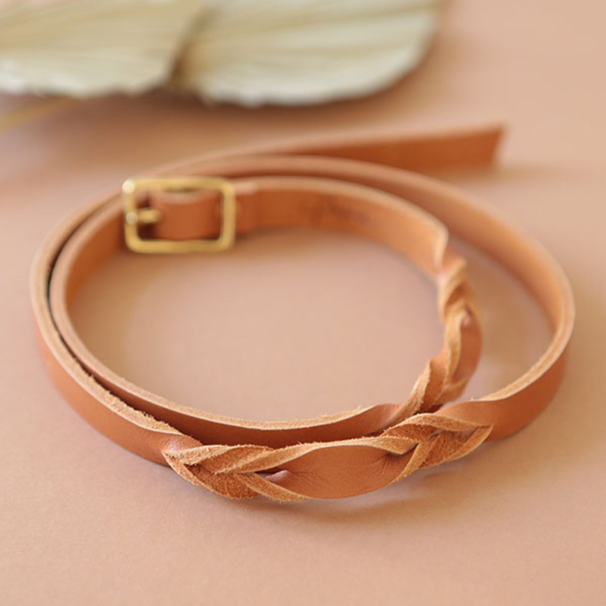 Geflochtener Ledergürtel in hellem Braun mit goldener Schnalle auf einem beigen Hintergrund.