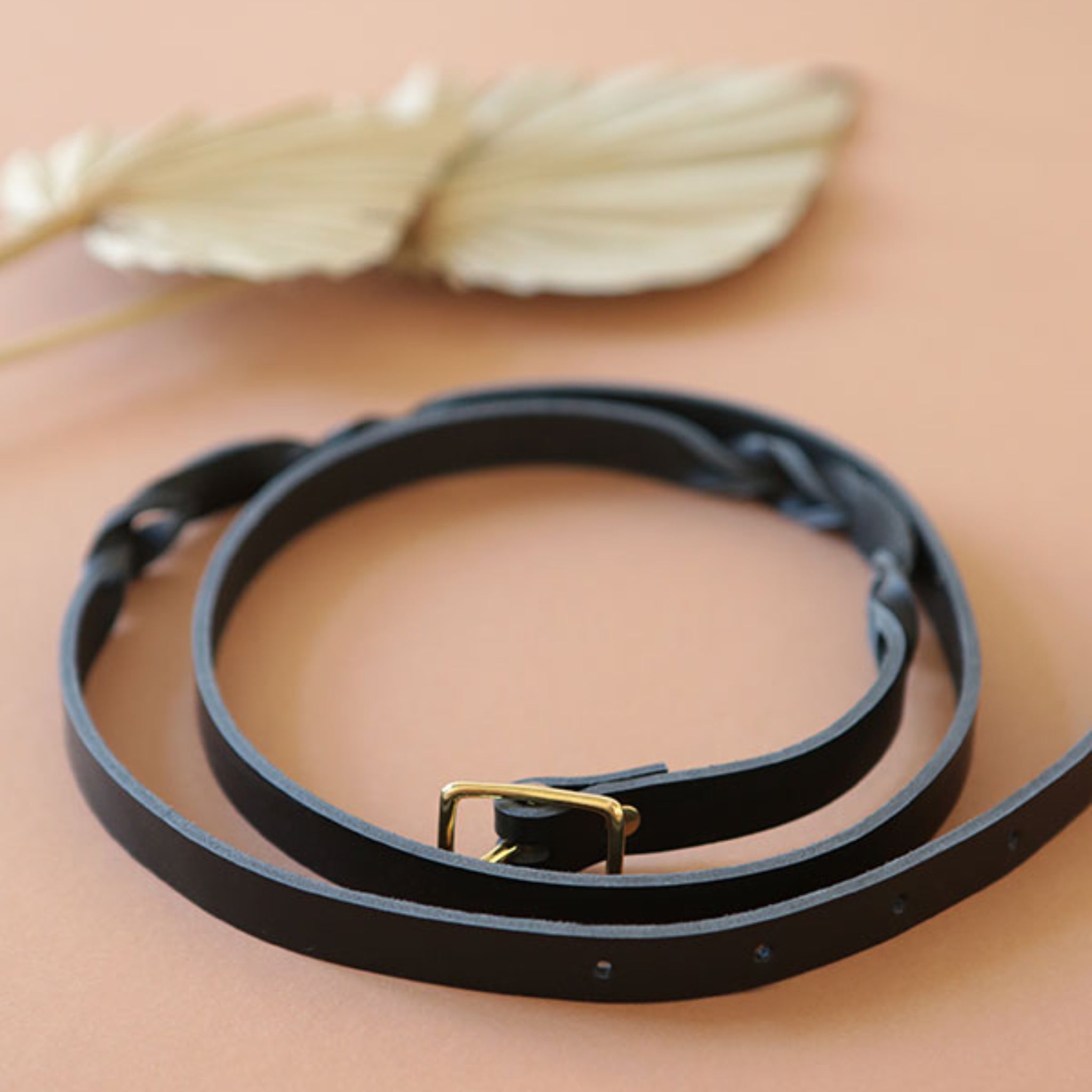 Schwarzer Ledergürtel mit goldener Schnalle liegt auf einem hellbraunen Hintergrund neben einem getrockneten Blatt.