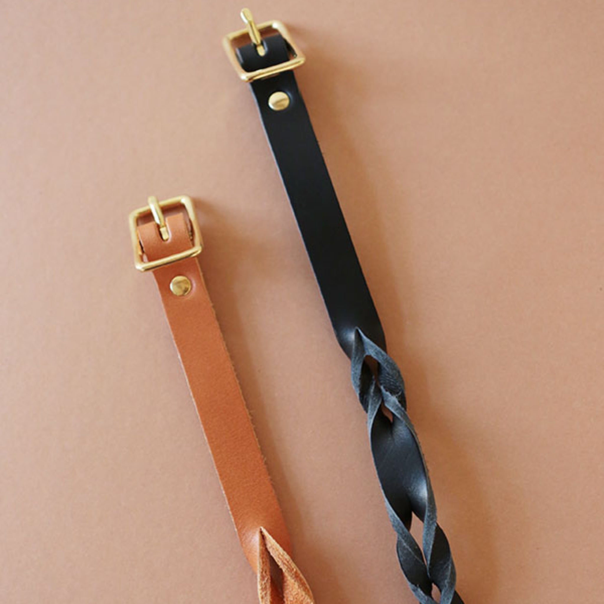 Zwei geflochtene Ledergürtel mit goldenen Schnallen, einer in Braun und einer in Schwarz, auf einem hellbraunen Hintergrund.