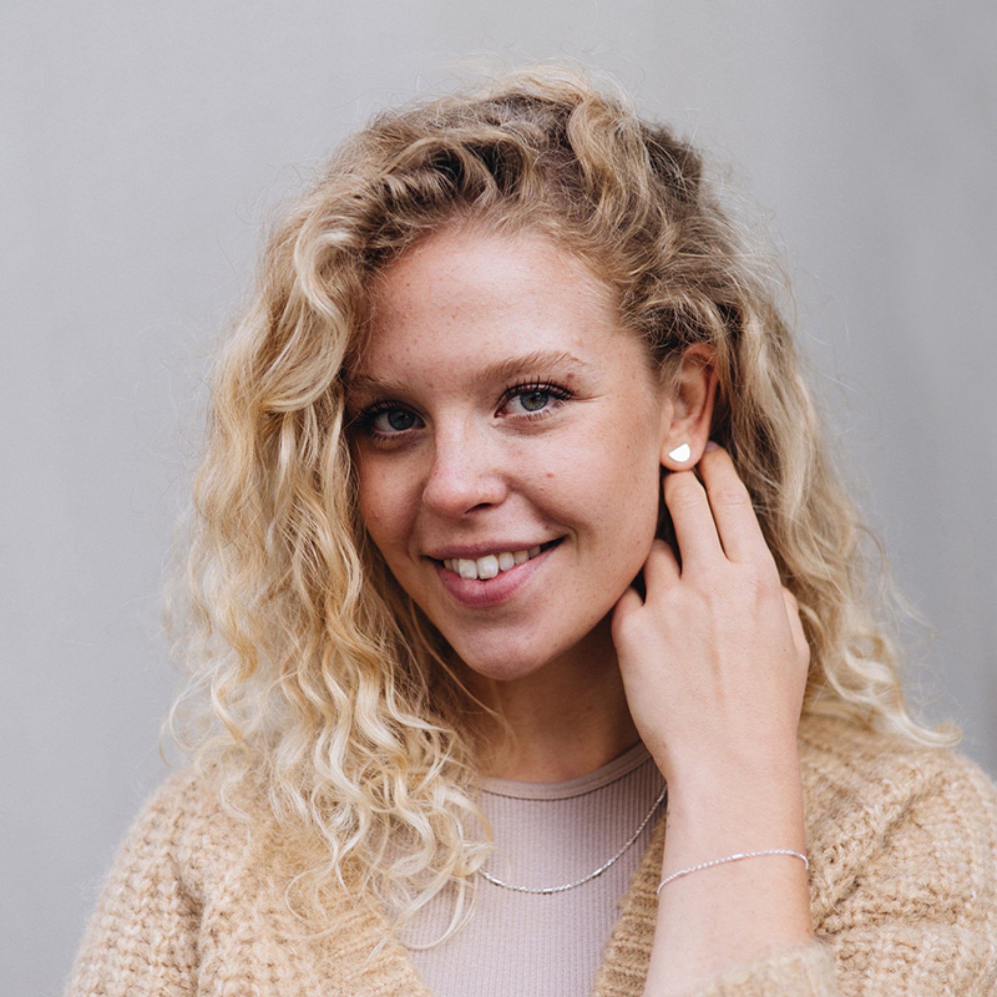 Eine junge Frau mit lockigem blondem Haar lächelt in die Kamera. Sie trägt einen beigen Pullover, ein helles Oberteil, eine dünne Halskette und ein Armband, wobei eine Hand ihr Ohr berührt. Der Hintergrund ist schlicht und in hellen Farben gehalten.