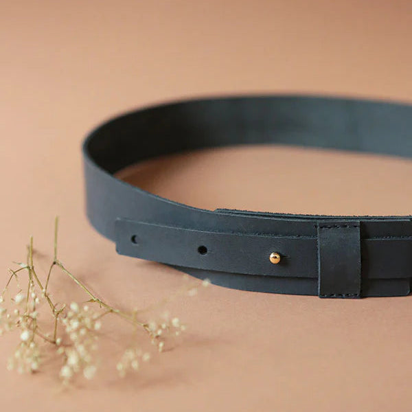 Ein schwarzer Ledergürtel mit einer goldenen Niete liegt auf einer hellbraunen Fläche neben einem kleinen Zweig mit zarten weißen Trockenblumen.