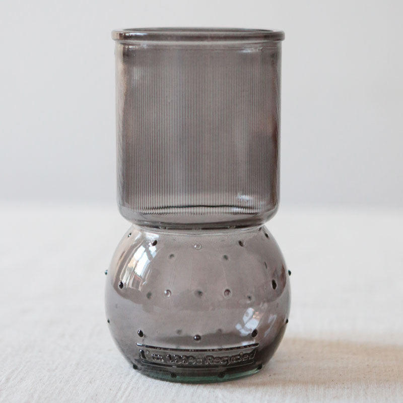 Ein modernes, transparentes, graues Trinkglas mit zylindrischem Kopf und rundem, gewölbtem Boden. Das Glas steht auf einer weißen Fläche mit einem schlichten, hellen Hintergrund.