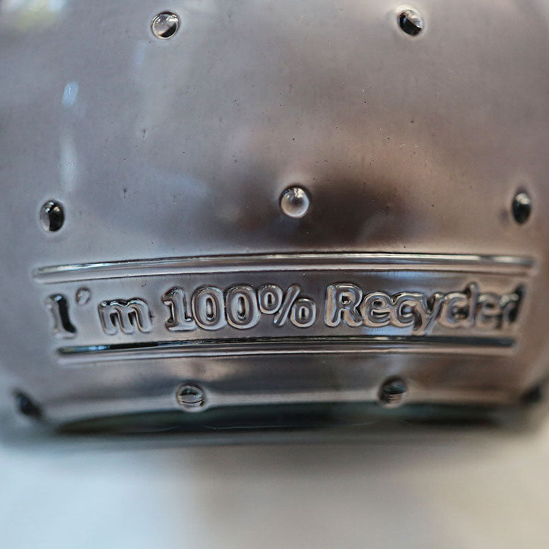 Nahaufnahme eines Glases mit geprägtem Text: "I'm 100% Recycled". Die Oberfläche weist kleine erhabene Punkte auf und der Gesamtton ist gräulich.