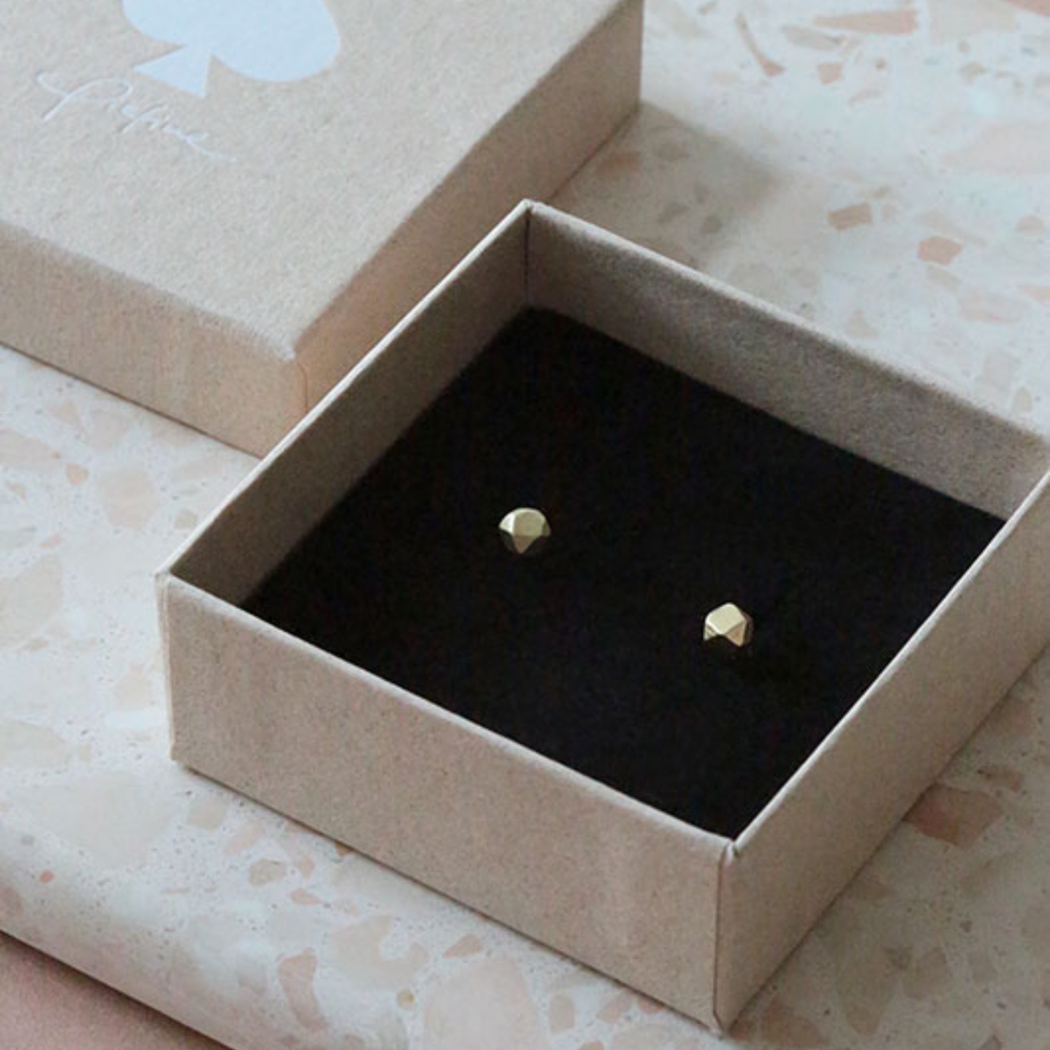 Ein kleines, offenes Schmuckkästchen aus Pappe mit schwarzer Innenseite enthält ein Paar einfacher, runder goldener Ohrstecker. Das Kästchen liegt auf einer leicht strukturierten Oberfläche neben seinem passenden Deckel.