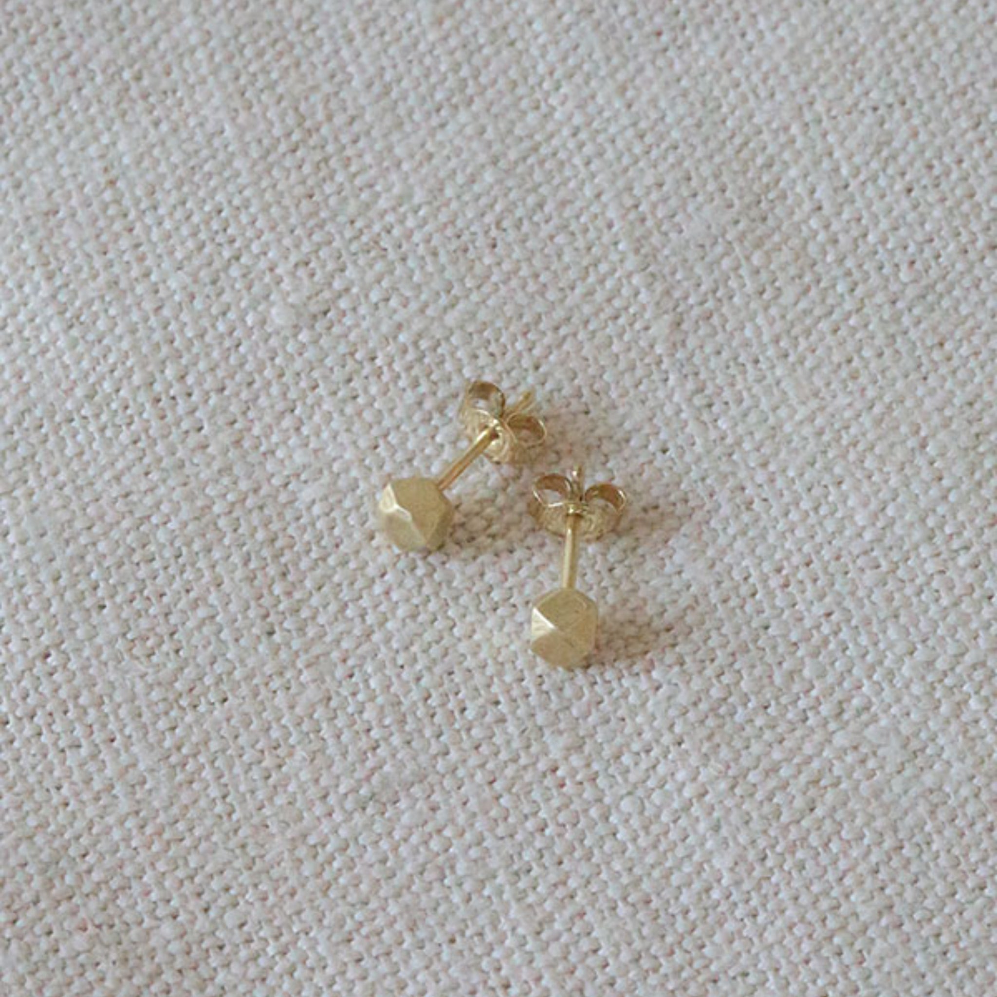 Ein Paar kleiner goldener Ohrstecker mit sechseckiger Vorderseite, die auf einer strukturierten beigen Stoffoberfläche angebracht sind.