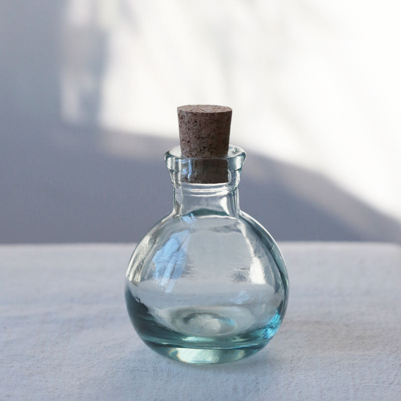 Eine runde, klare Glasflasche mit kurzem Hals und Korkenverschluss steht auf einer weißen Fläche, mit weichem natürlichem Licht und subtilen Schatten im Hintergrund.