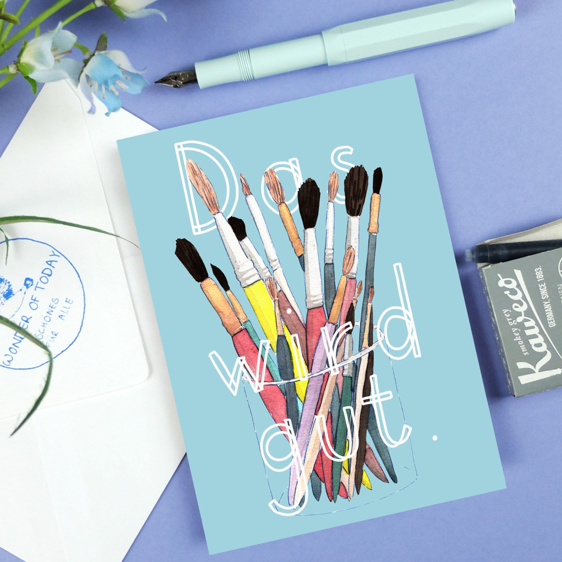 Postkarte mit Pinseln in einem Glas und dem Text „Das wird gut“ auf blauem Hintergrund, neben Füller und Umschlag.