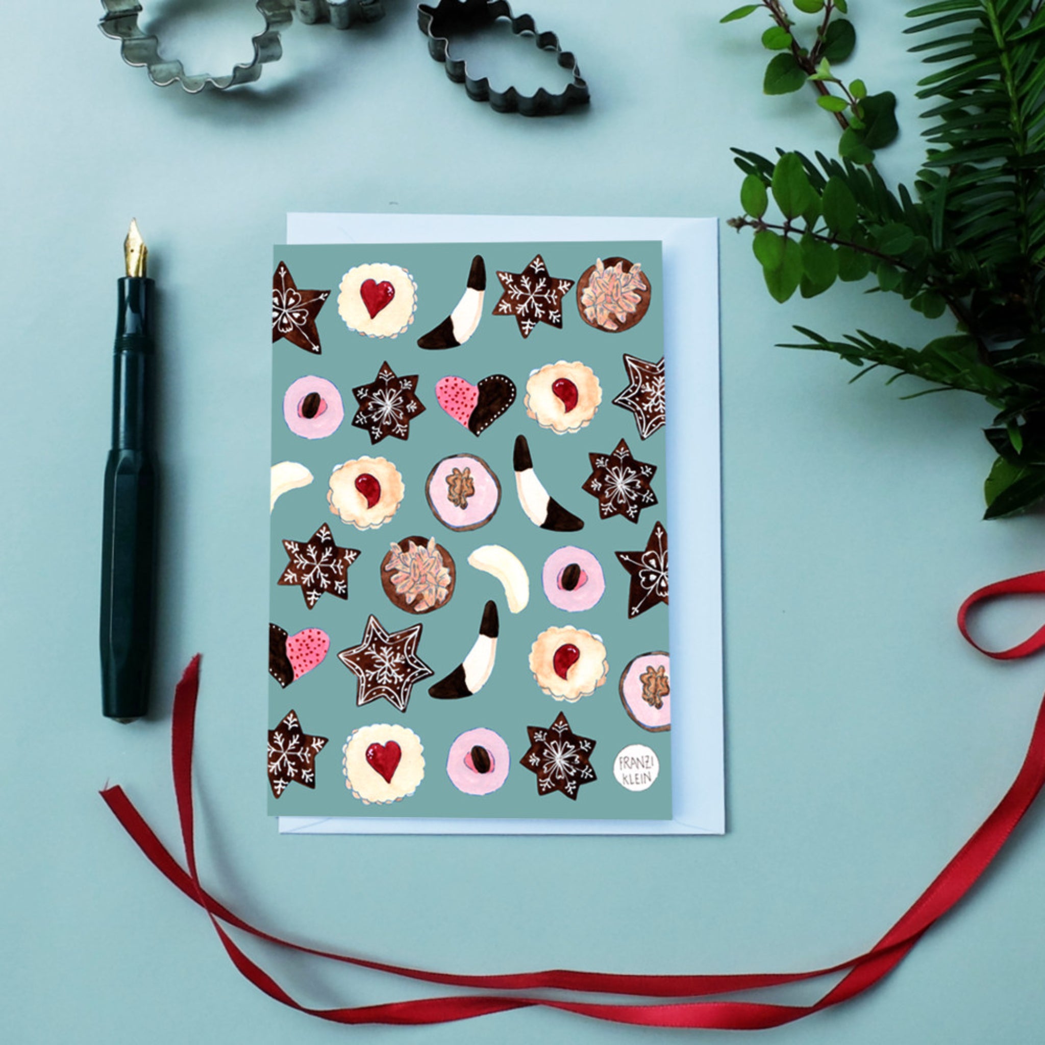 Grußkarte mit bunten Keksmustern auf blauem Hintergrund, neben einem Füllfederhalter, Ausstechformen und roten Band.