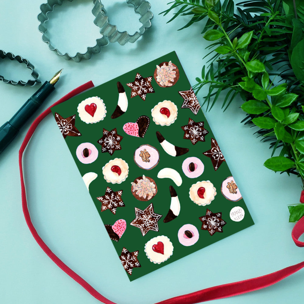 Grünes Notizbuch mit verschiedenen Gebäck- und Keksmustern, umgeben von Pflanzen und Backutensilien.