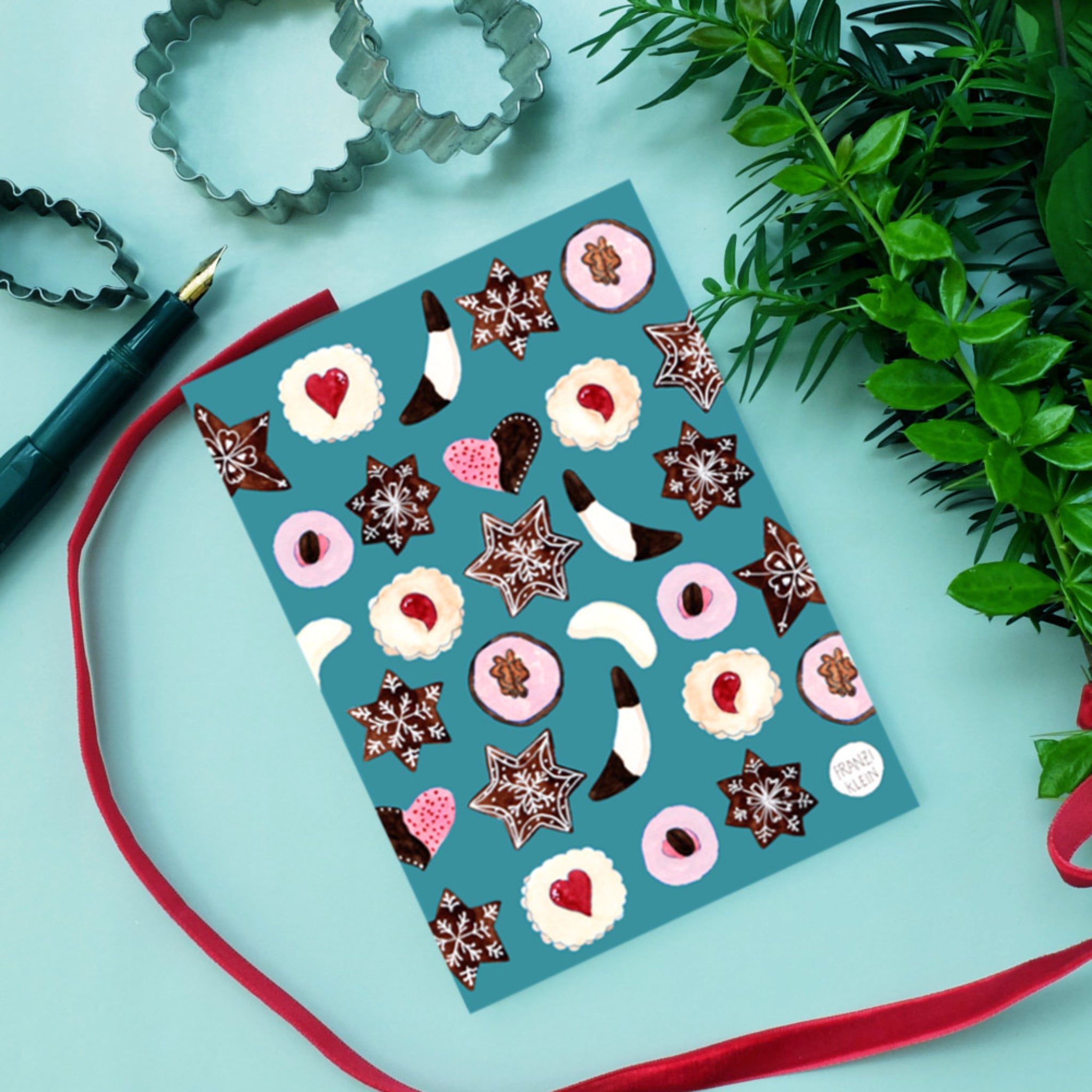 Karte mit Weihnachtsplätzchen auf blauem Hintergrund, umgeben von Backformen, Pflanzen und rotem Band.