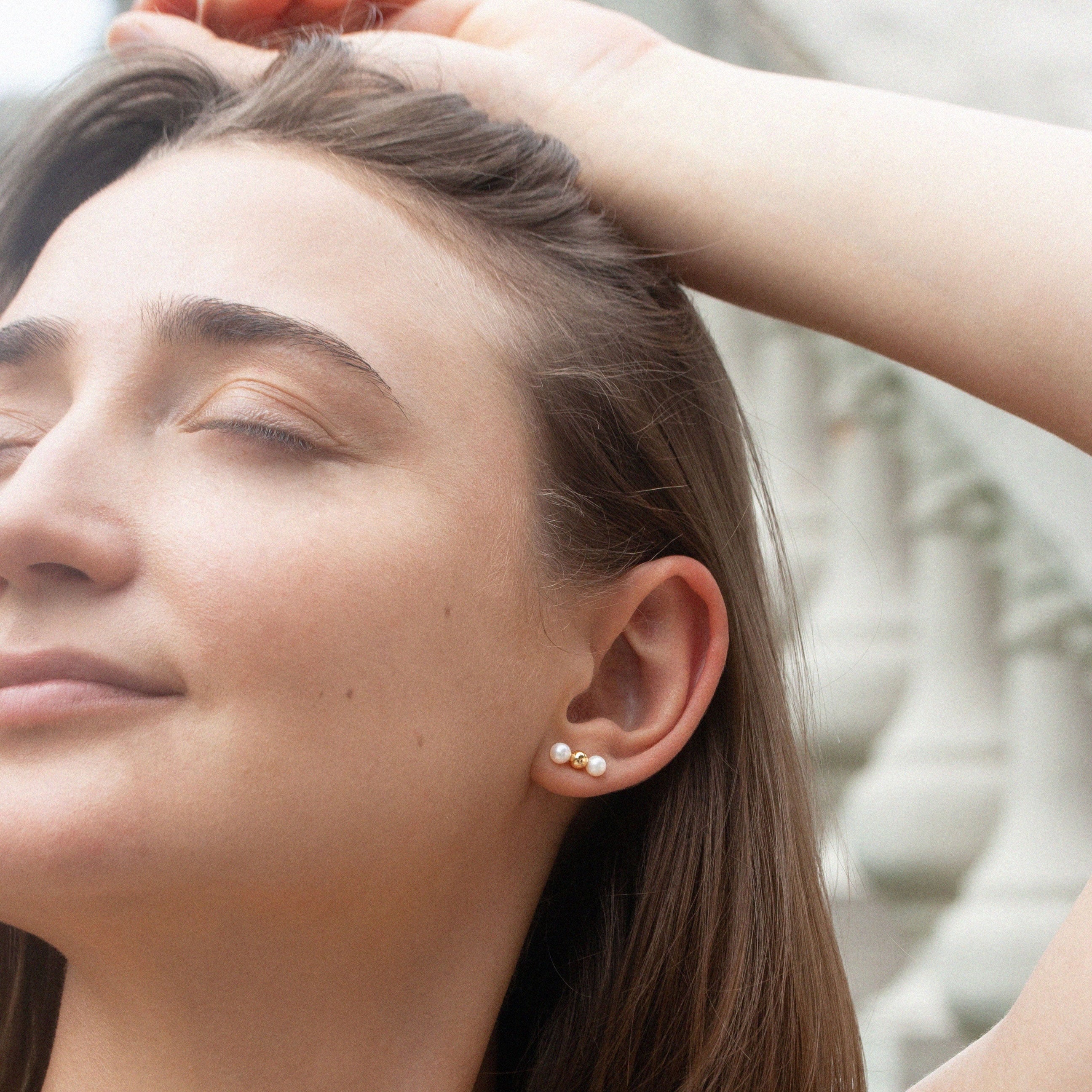 Frau mit geschlossenen Augen in Nahaufnahme, trägt kleinen goldenen Ohrring mit Perlen, entspannt im Freien.