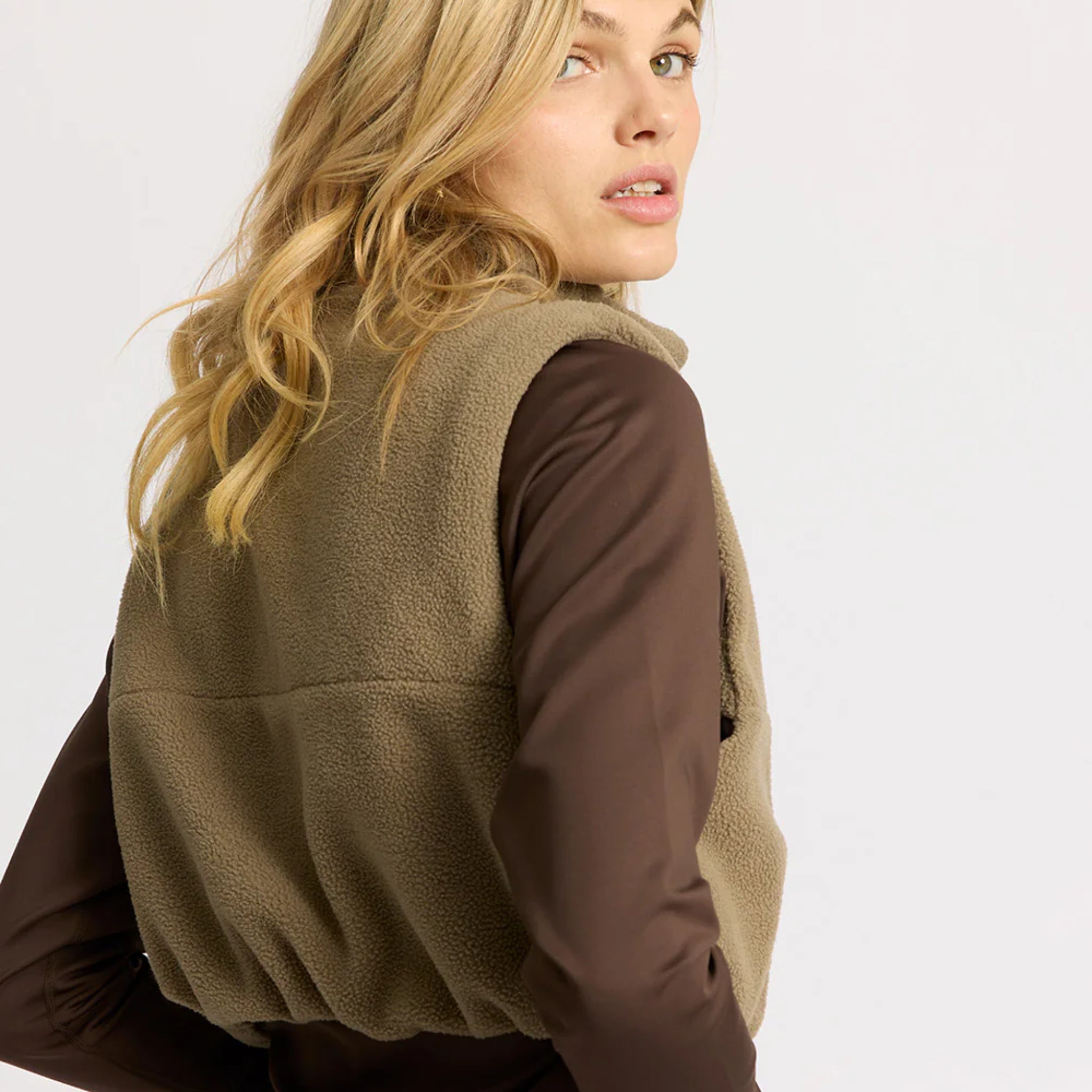 Eine Frau mit gewelltem blondem Haar trägt eine hellbraune Fleece-Weste über einem langärmeligen braunen Hemd. Sie posiert mit dem Rücken zugewandt und schaut mit dem Kopf über ihre Schulter vor einem schlichten hellen Hintergrund.